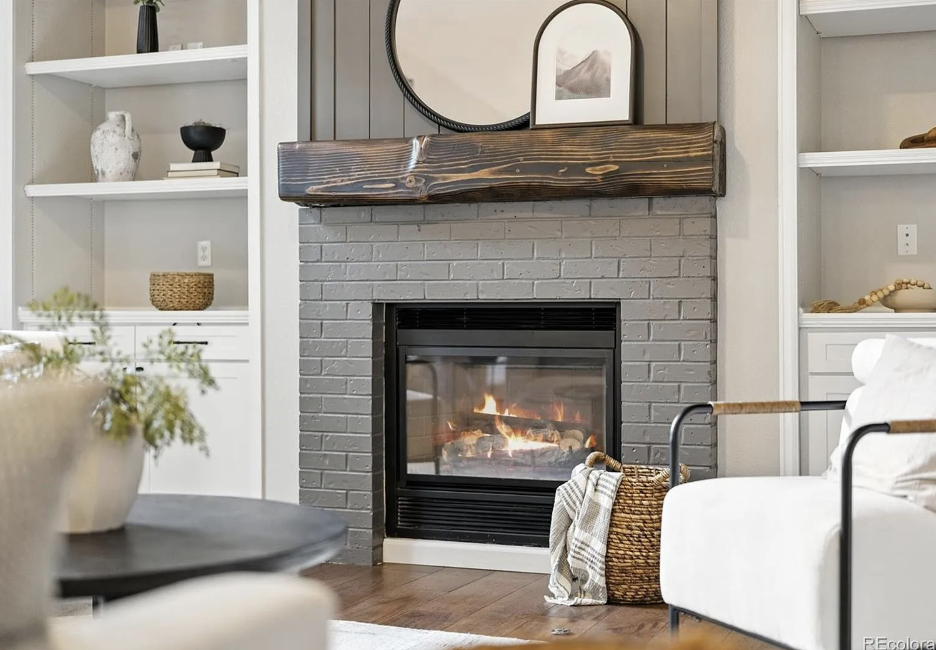 Living room with a fireplace, white couch, shelves with decorative items, and a round mirror on the wall.