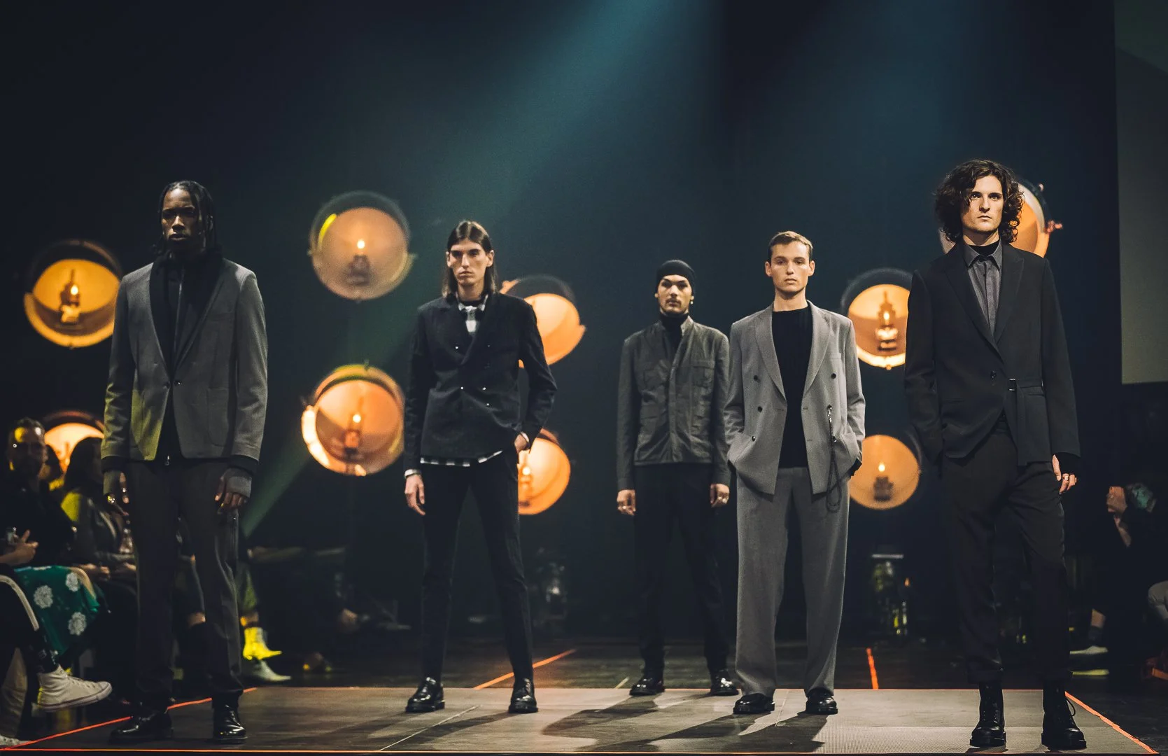 Cinq mannequins masculins et féminins défilant sur un podium lors d'un défilé de mode avec un éclairage chaud en fond