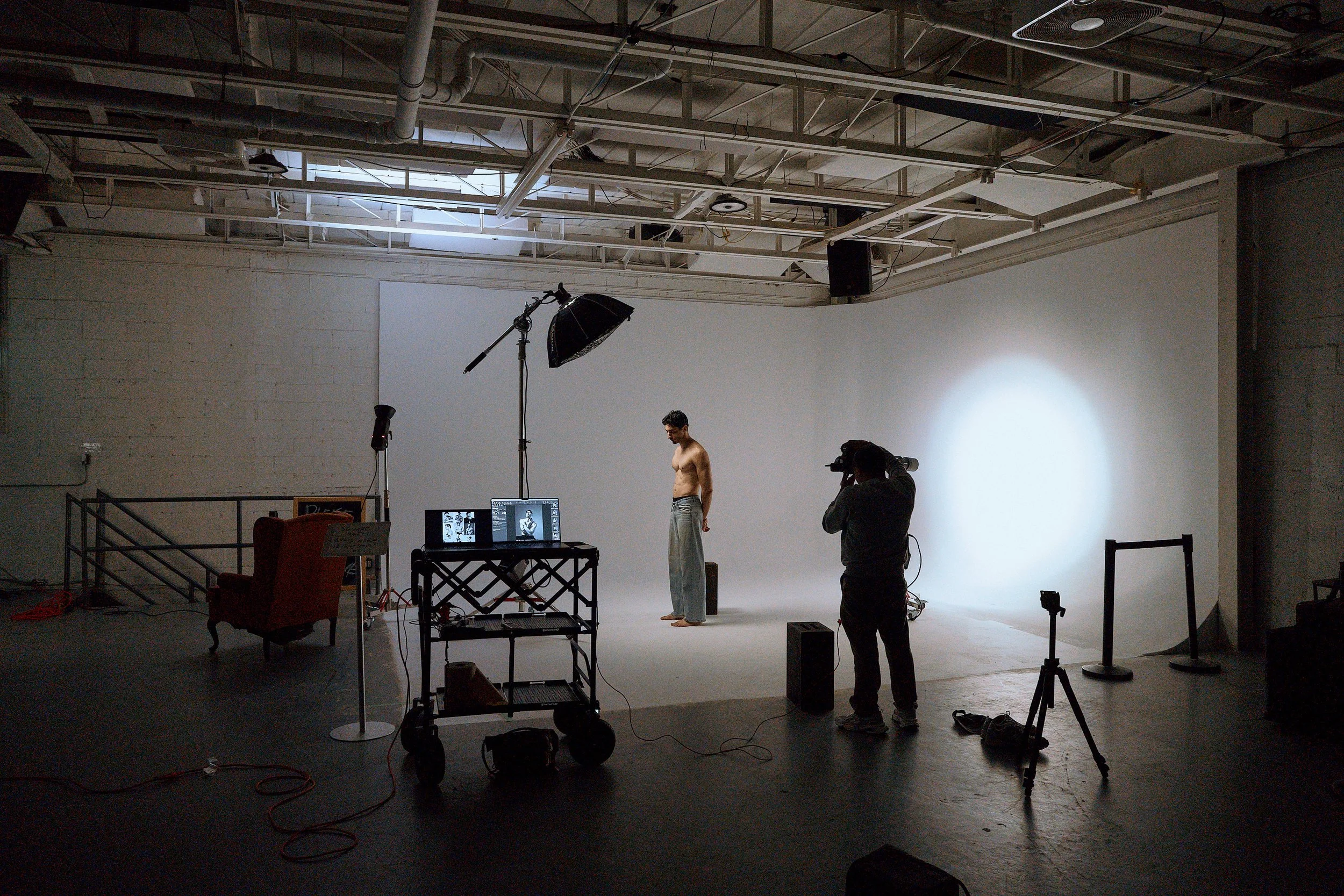 Un homme nu est en train de poser dans un studio de photographie avec éclairage professionnel, un photographe le prend en photo, avec un fond blanc lumineux.