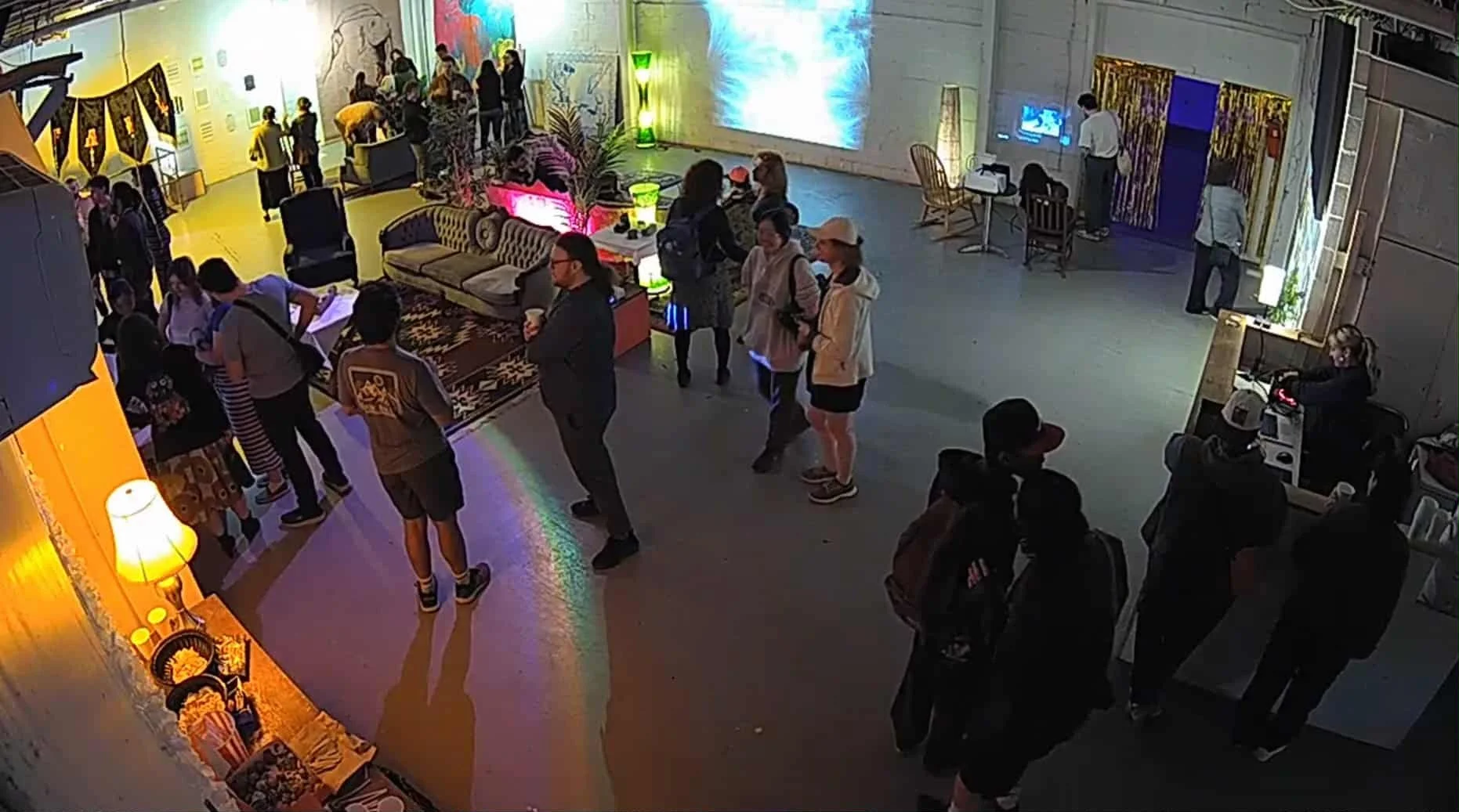 Un groupe de personnes dans un espace intérieur coloré, avec des meubles vintage, un écran lumineux, une petite scène avec un DJ, et des décorations lumineuses.