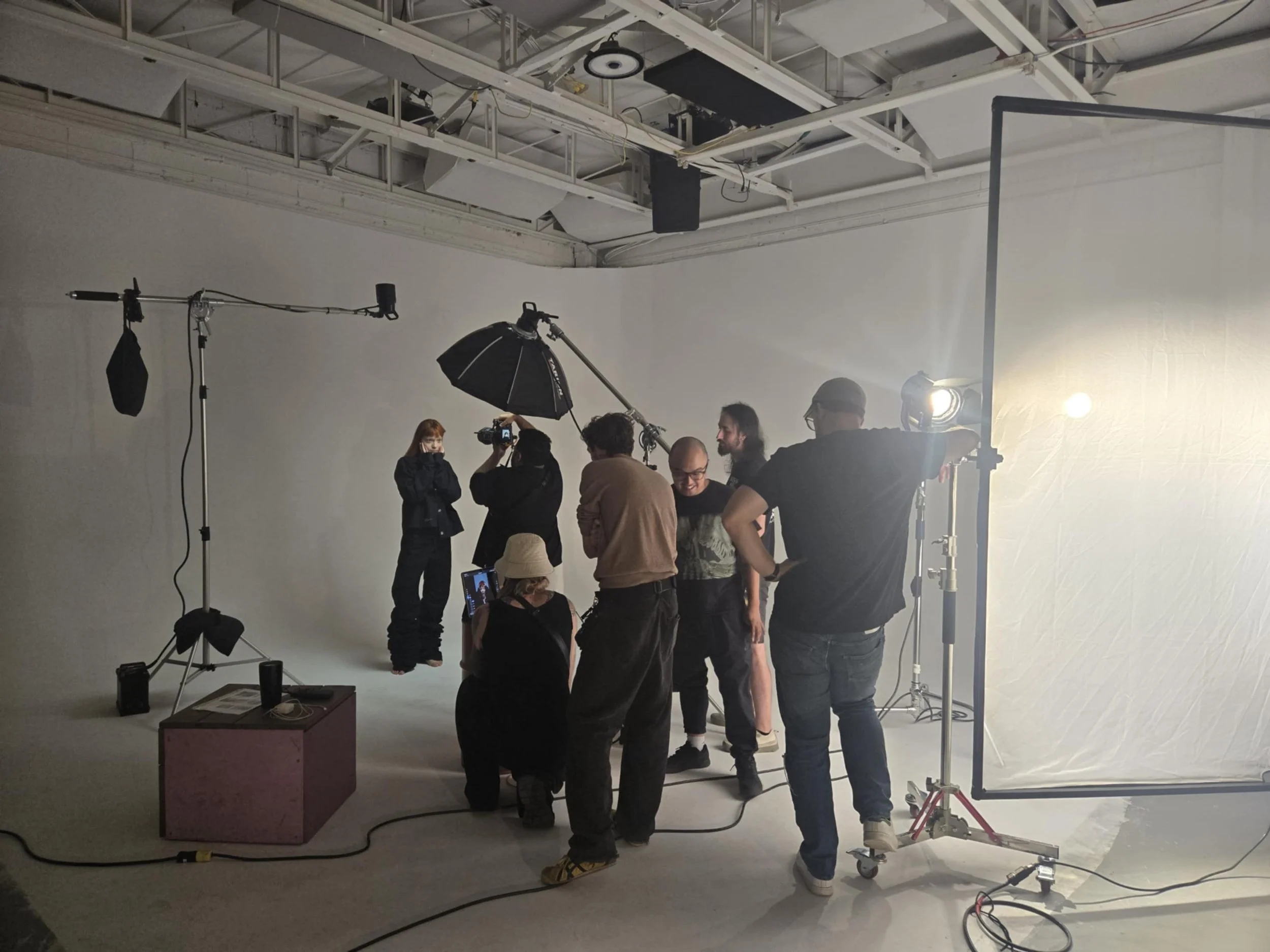 Une équipe de production en studio photo ou vidéo, avec un fond blanc, des équipements d’éclairage, un umbrella, un diffuseur et plusieurs personnes en train de préparer la prise de vue.