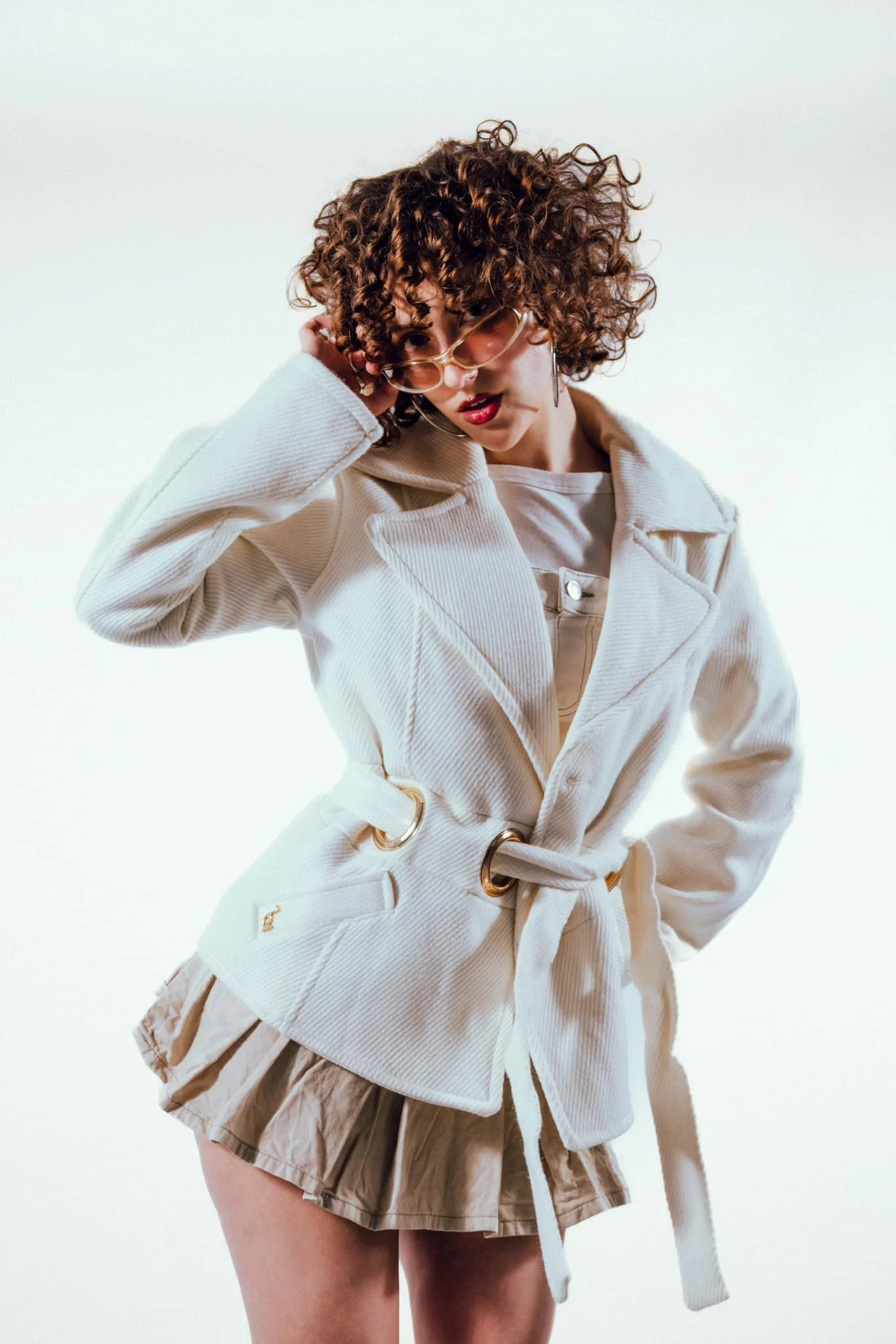 Femme aux cheveux bouclés portant des lunettes, une veste blanche et une jupe beige