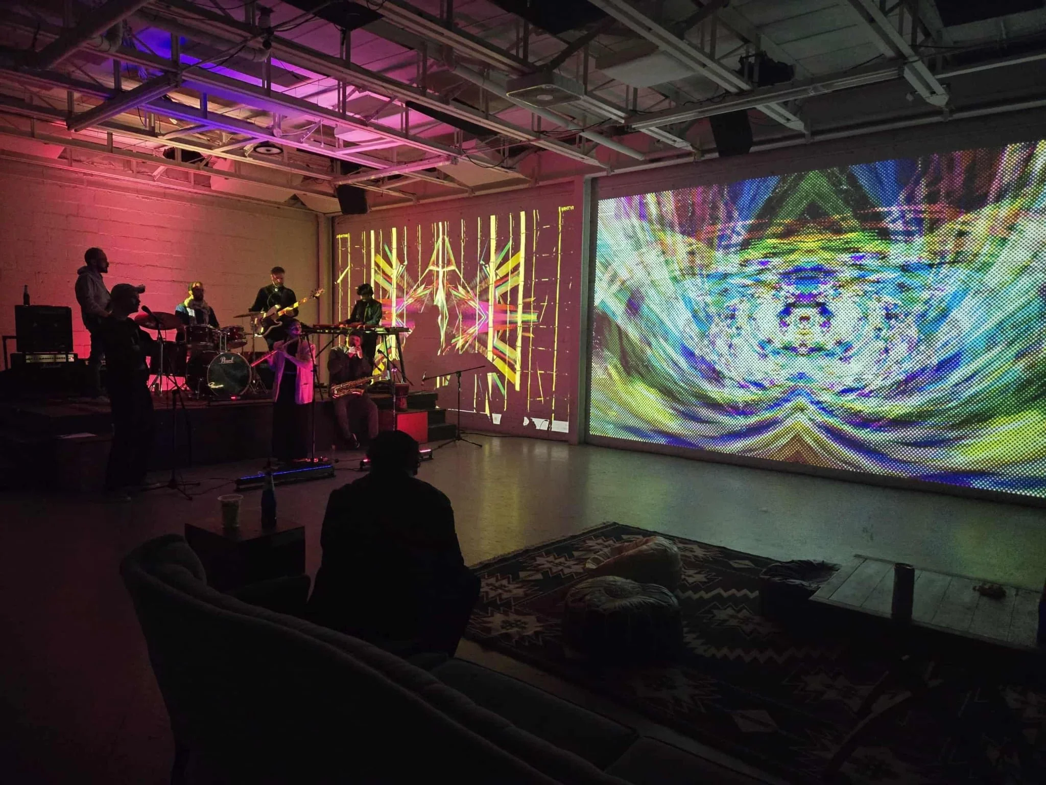 Un groupe de musiciens joue sur scène dans une pièce avec des murs blancs, sous des lumières colorées violettes et roses, avec un grand écran LED affichant des motifs géométriques et psychédéliques.