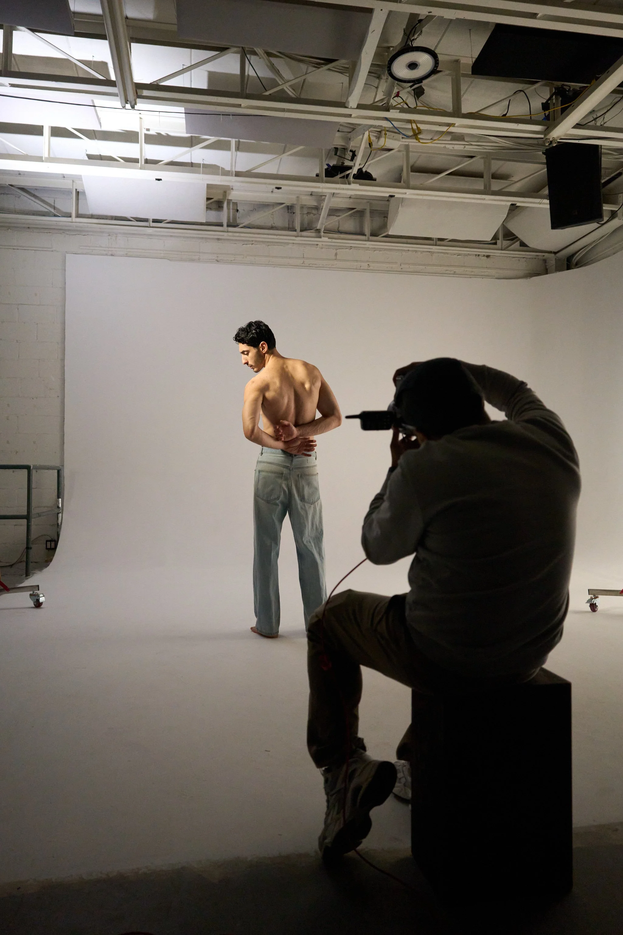Un homme nu en train de poser pour une séance photo dans un studio avec un fond blanc, tandis qu'un photographe l'observe et prend des clichés.