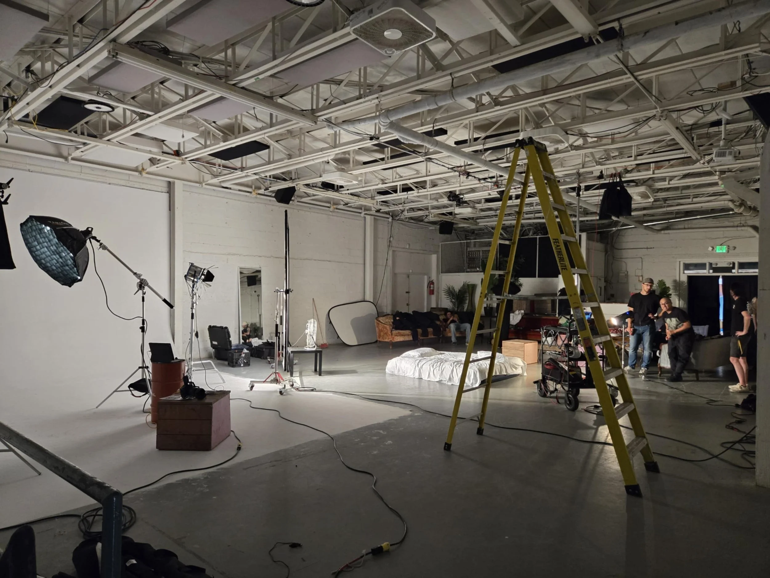 Un studio de tournage avec un fond blanc, un éclairage professionnel, un lit au sol, des meubles et des personnes en train de préparer le tournage.