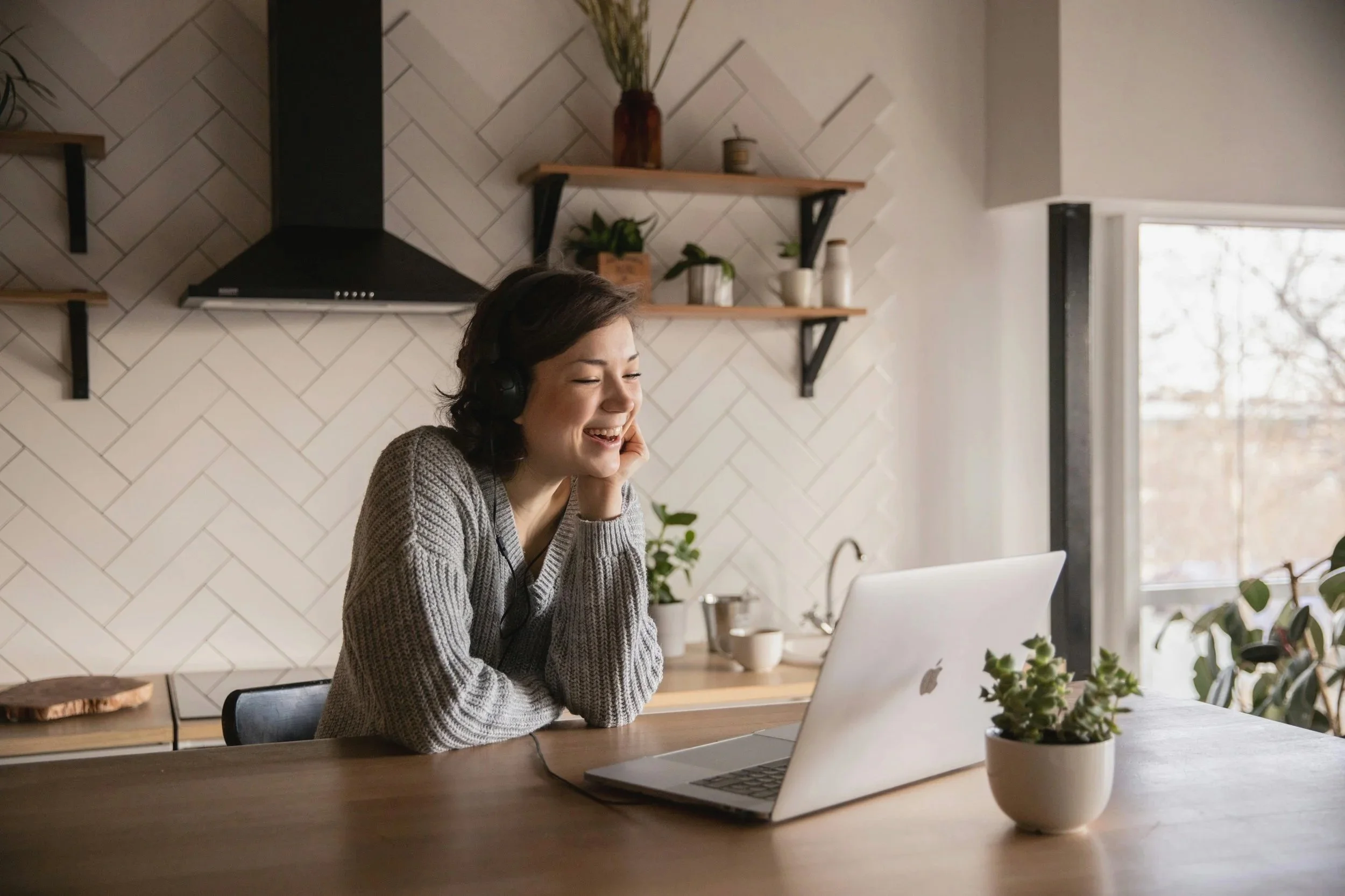 A woman sitting at a wooden table in a bright kitchen, smiling while talking on a video call on her laptop, with plants and kitchen decor in the background.