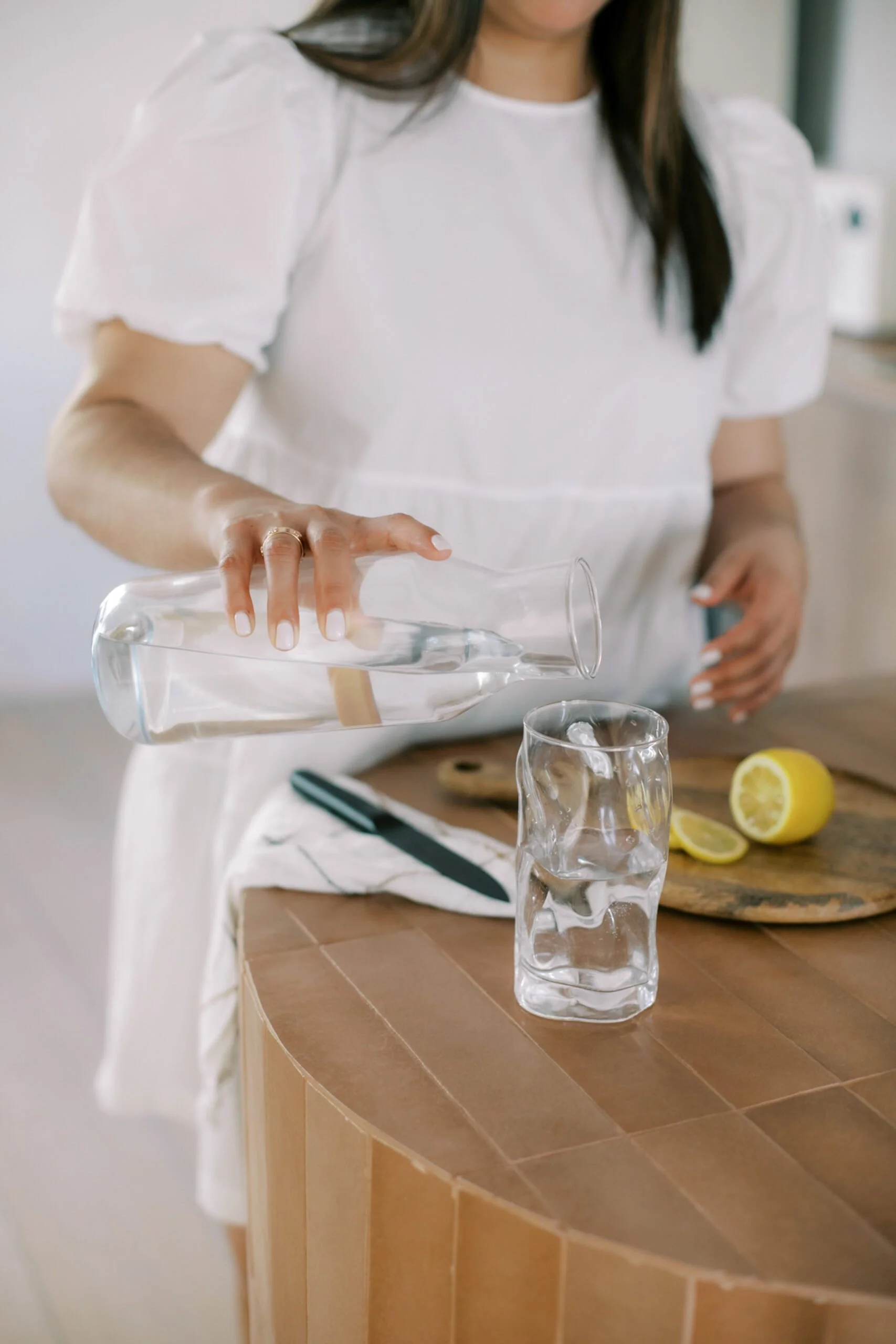 A woman pouring water into a glass with ice, on a wooden table that has a lemon and a knife.