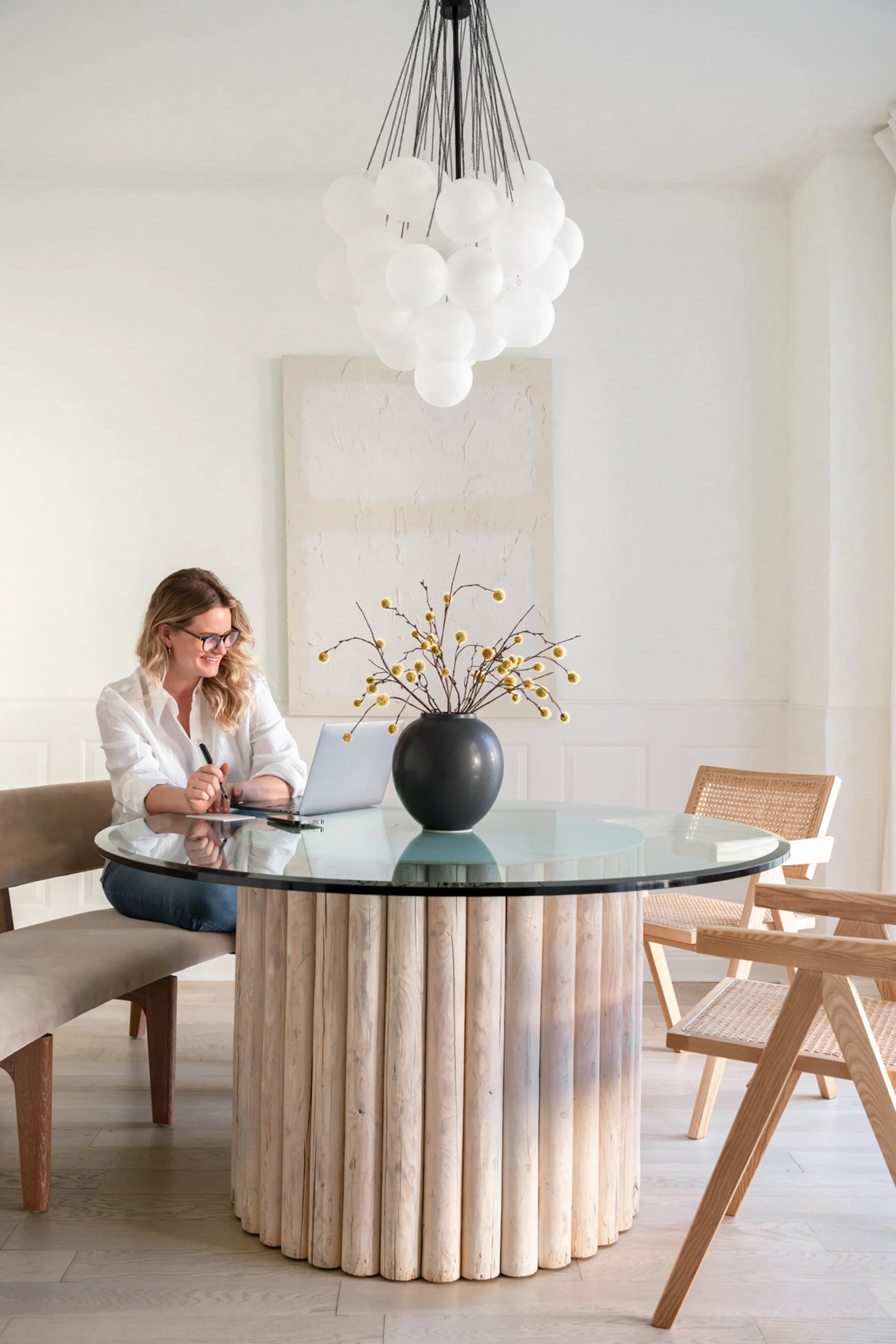 Woman sitting at a round glass-top table with a laptop, engaged in work, in a brightly lit, minimalist dining or meeting room with neutral decor.