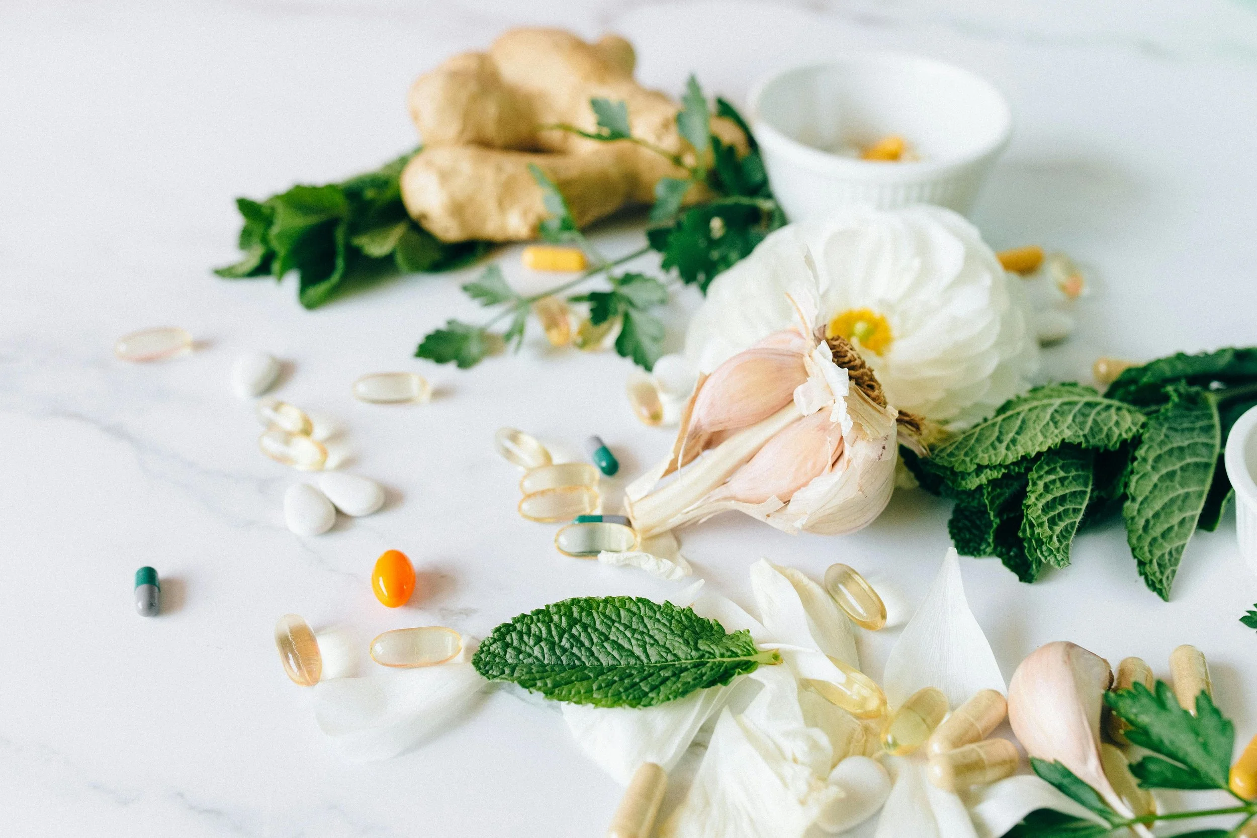 Assorted herbs, garlic, flowers, and pills scattered on a white surface.