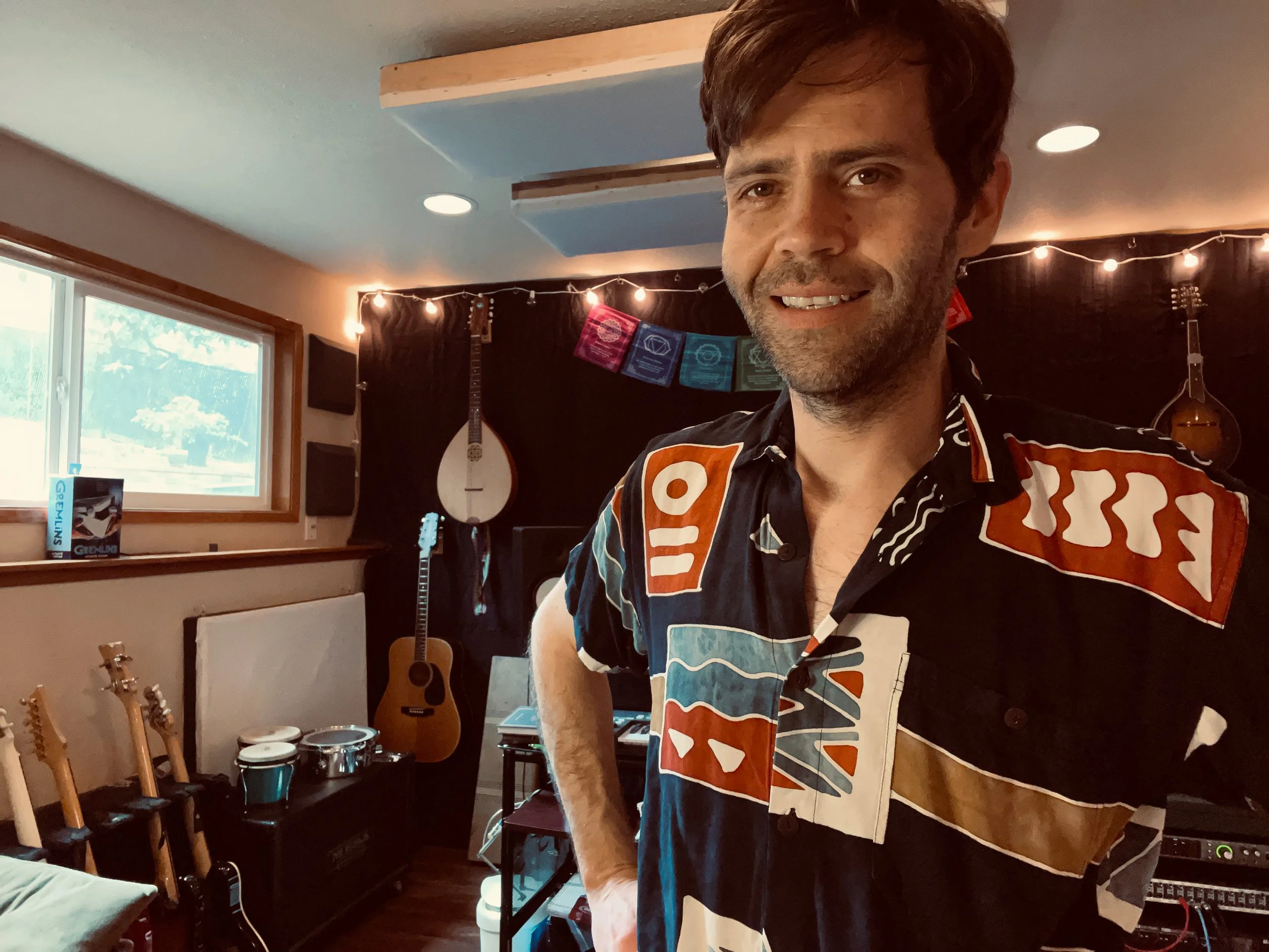 Man with colorful shirt in a music studio with guitars, drums, and string lights.