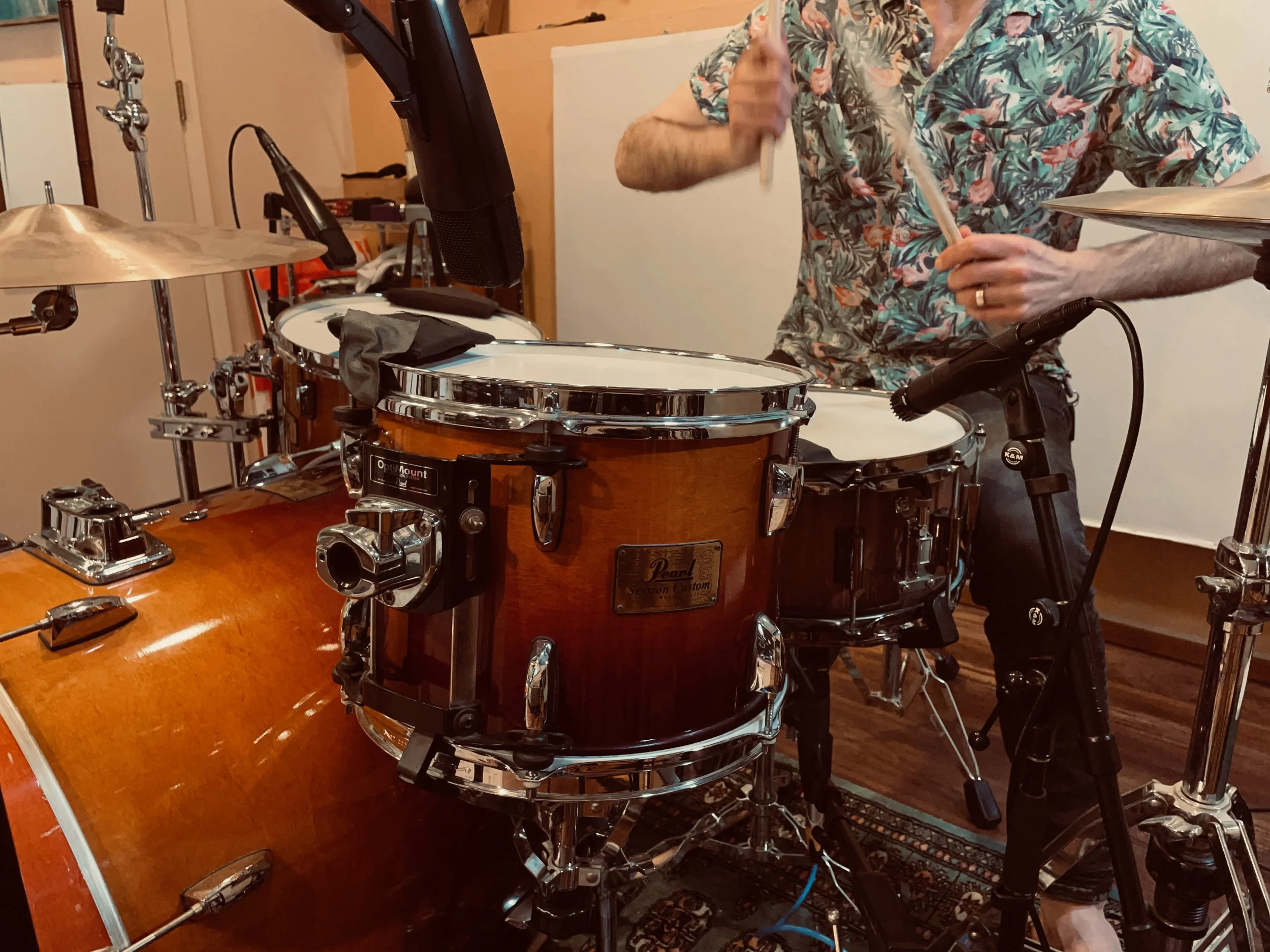 Person playing a drum set with microphones, wearing a floral shirt, in a music studio setting.