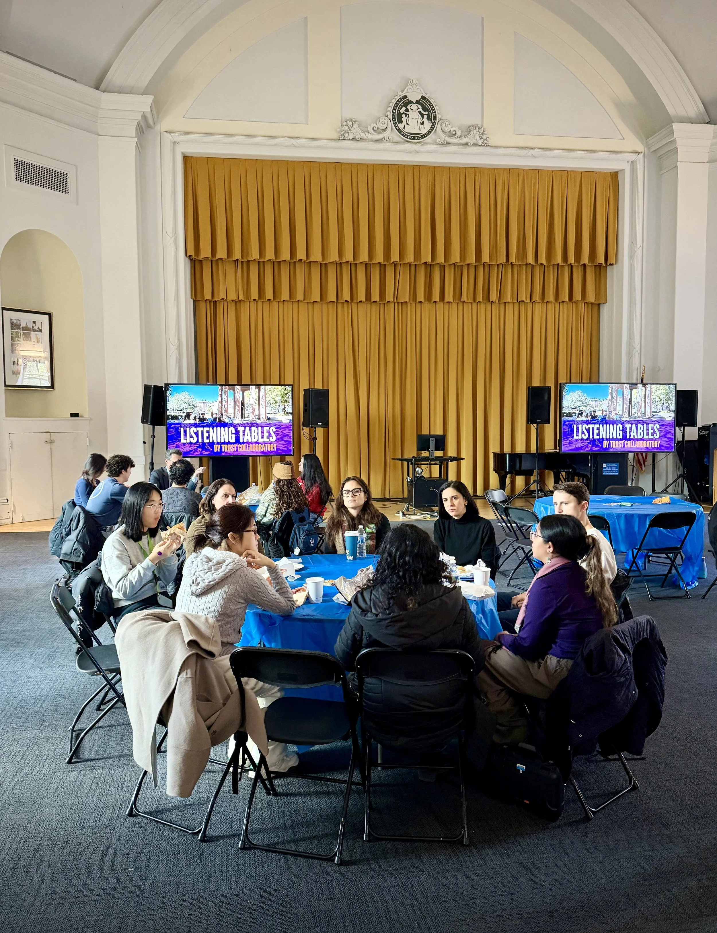 People sitting around a round table at a conference or workshop, with a large stage and two screens in the background displaying 'Listening Tables' by Trust Collaboratory.