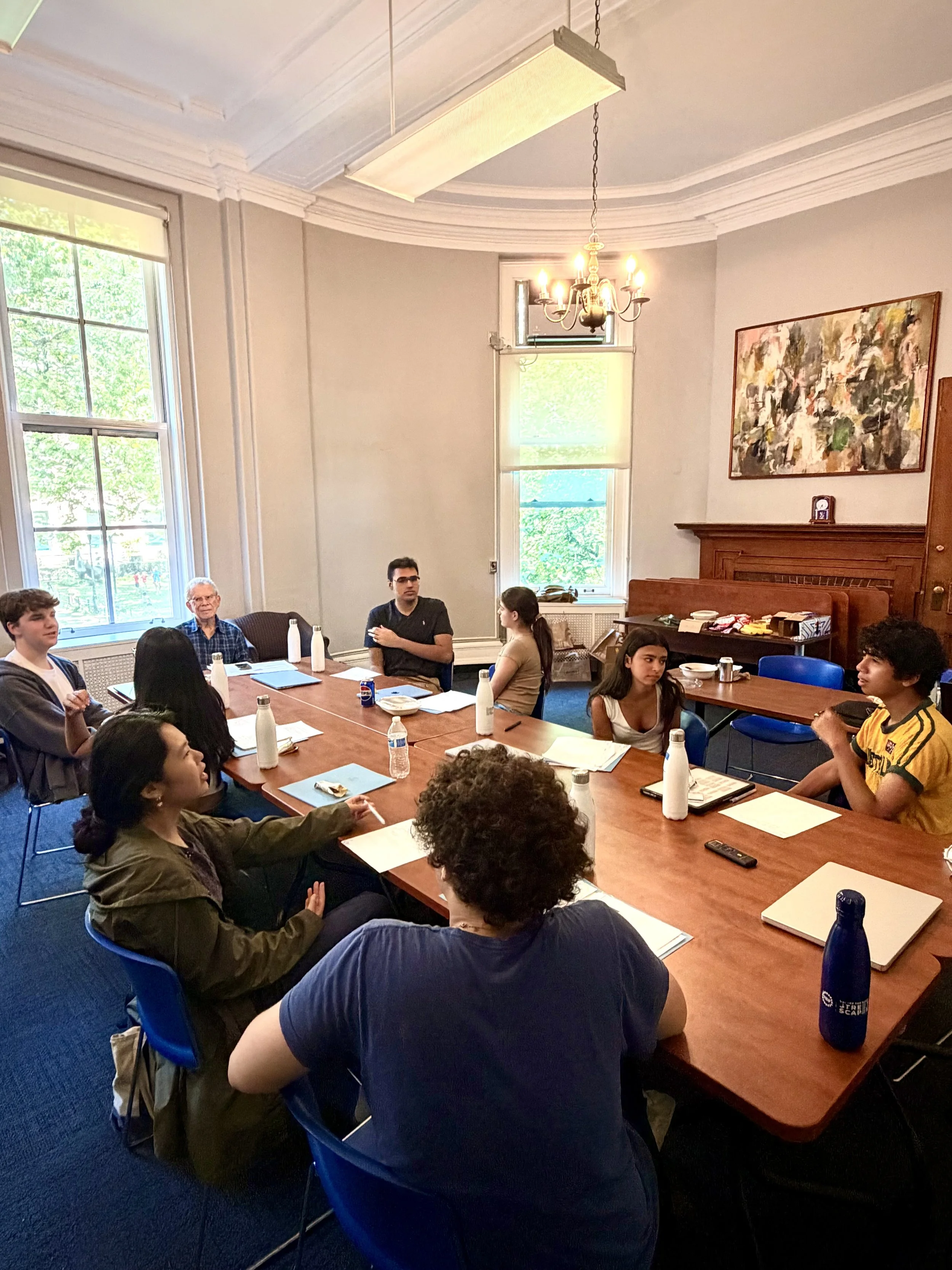 A group of diverse young adults and an older man sit around a large wooden table in a well-lit room with high ceilings, large windows, and a chandelier. The table has water bottles, papers, and notebooks. People appear to be engaged in a discussion or meeting.