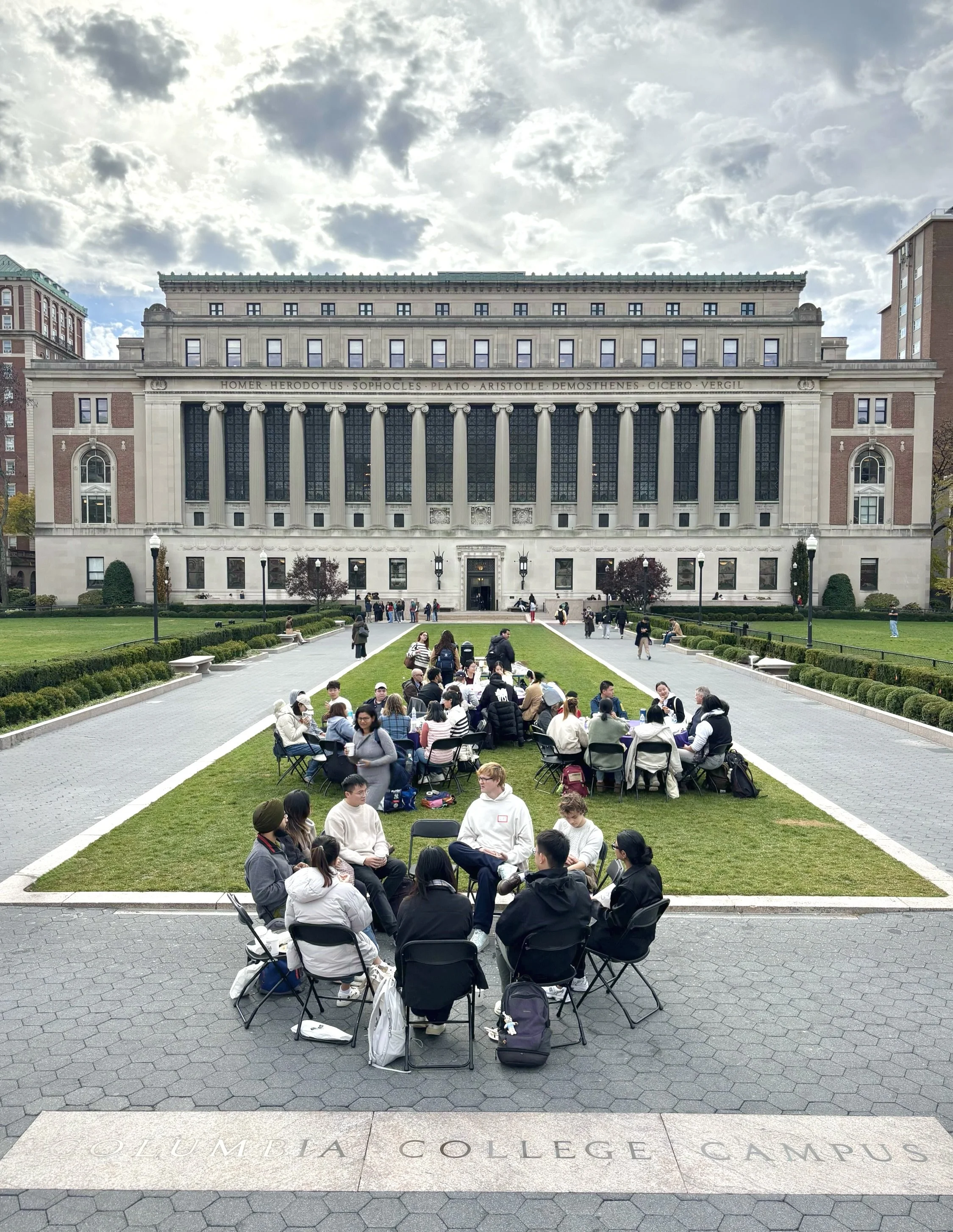 Students sitting and standing on the grassy area and sidewalk of Columbia College Campus, engaging in group discussions and activities under a partly cloudy sky, with a large historic building in the background.