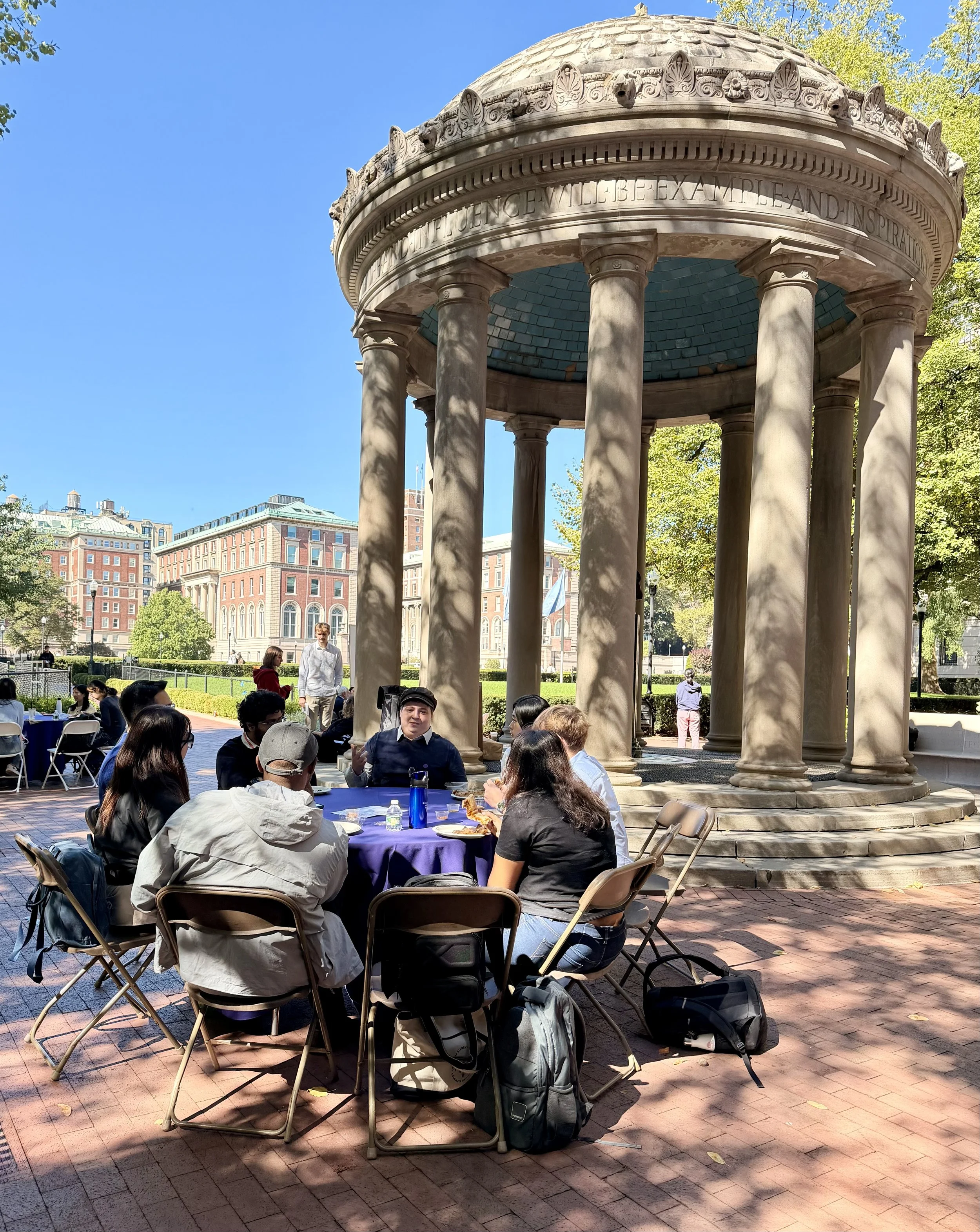 People seated around a table with food, enjoying an outdoor gathering near a historic bandstand in a city park.