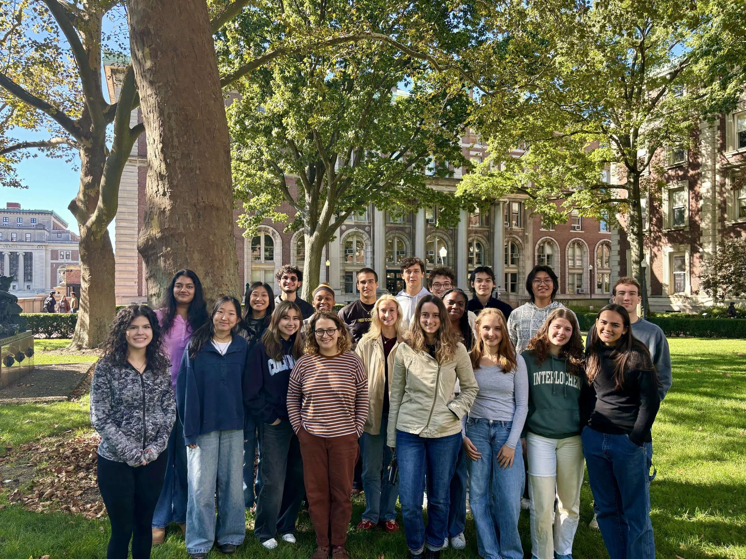 Group of young adults standing together outdoors in a park with green trees and buildings in the background, smiling and posing for a photo.
