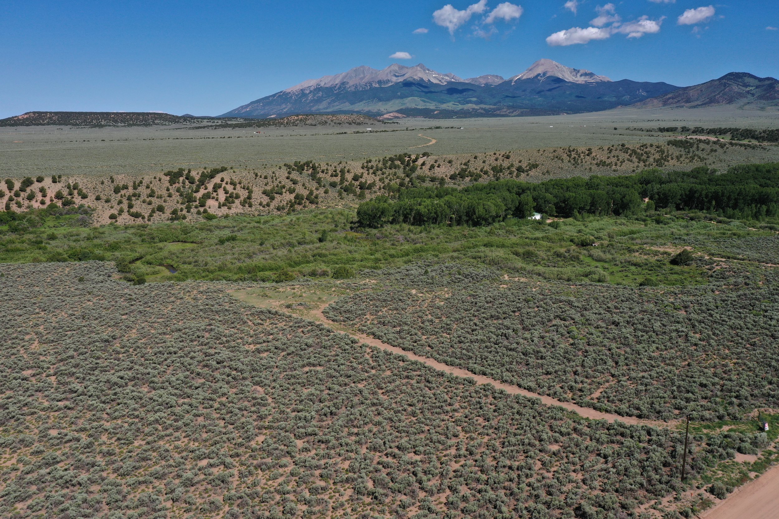 A vast landscape featuring a dirt road cutting through green bushes and trees, with rolling hills leading to distant mountains under a bright blue sky with scattered clouds.