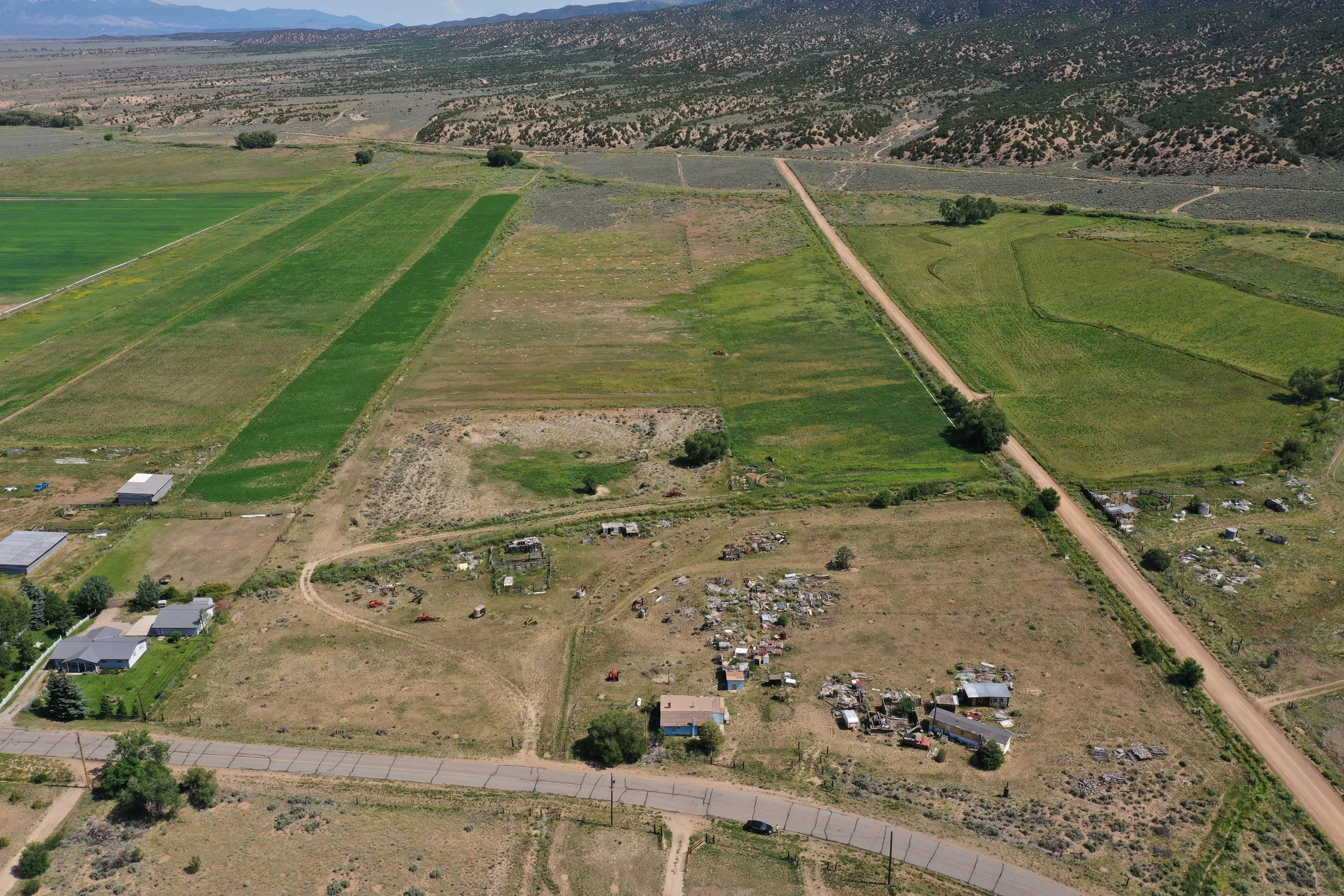 Aerial view of farmland and a small residential area with some houses, roads, and patches of green fields, set against a backdrop of hills or mountains.