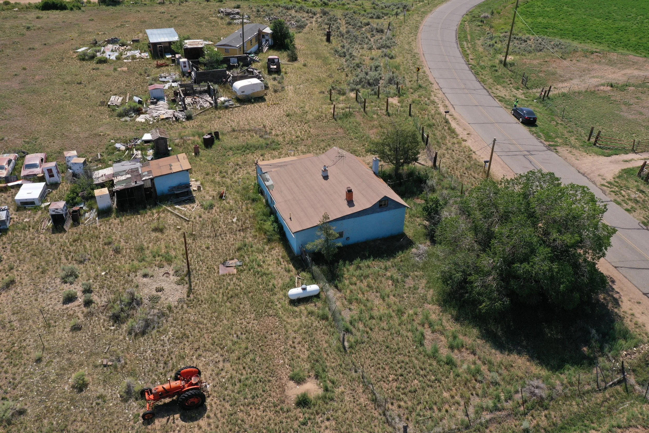 An aerial view of a rural property with a small blue house, various debris and old vehicles scattered around, surrounded by open grassland and a winding road nearby.