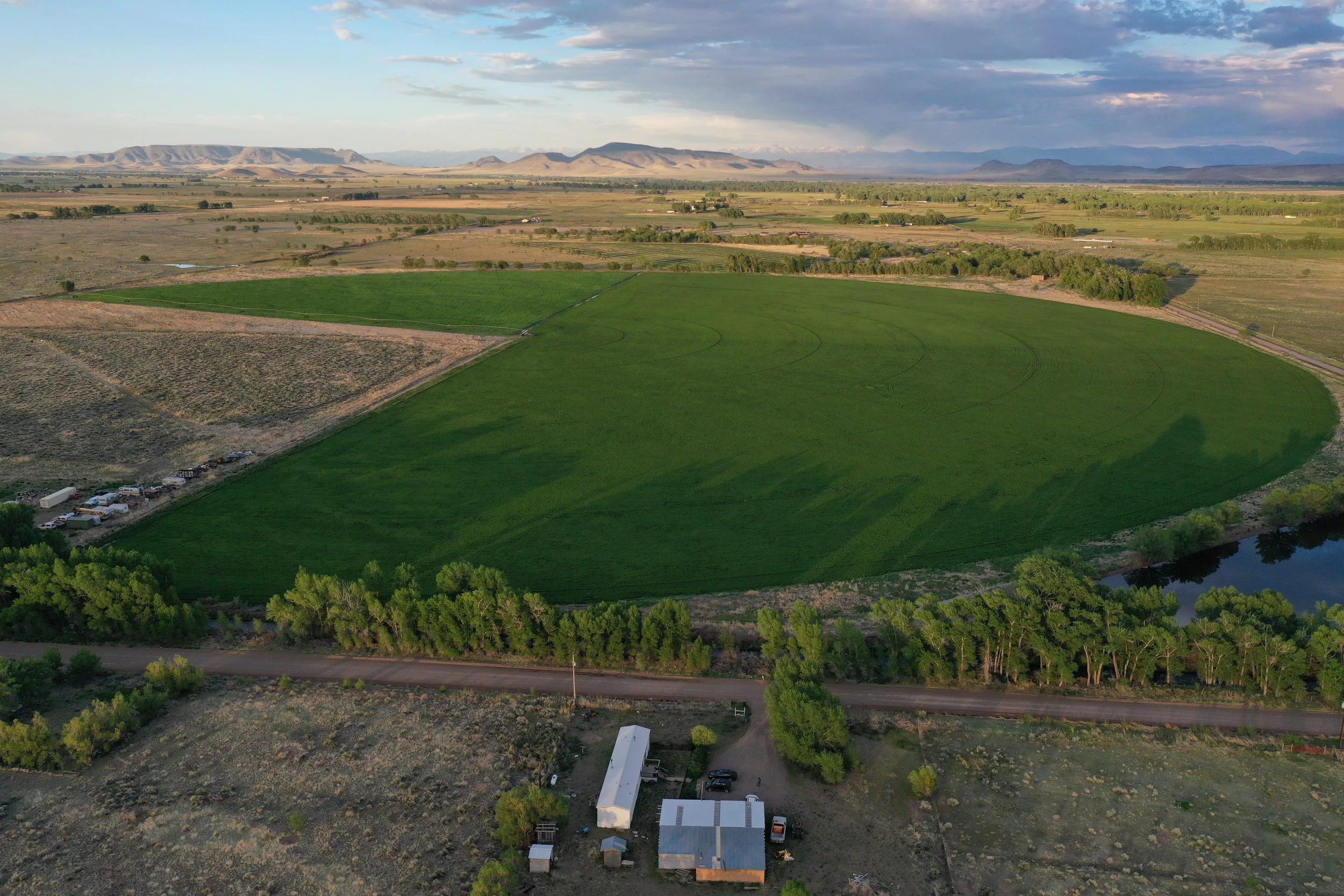 Aerial view of a rural landscape with green fields, trees, a river, and distant mountains under a partly cloudy sky.