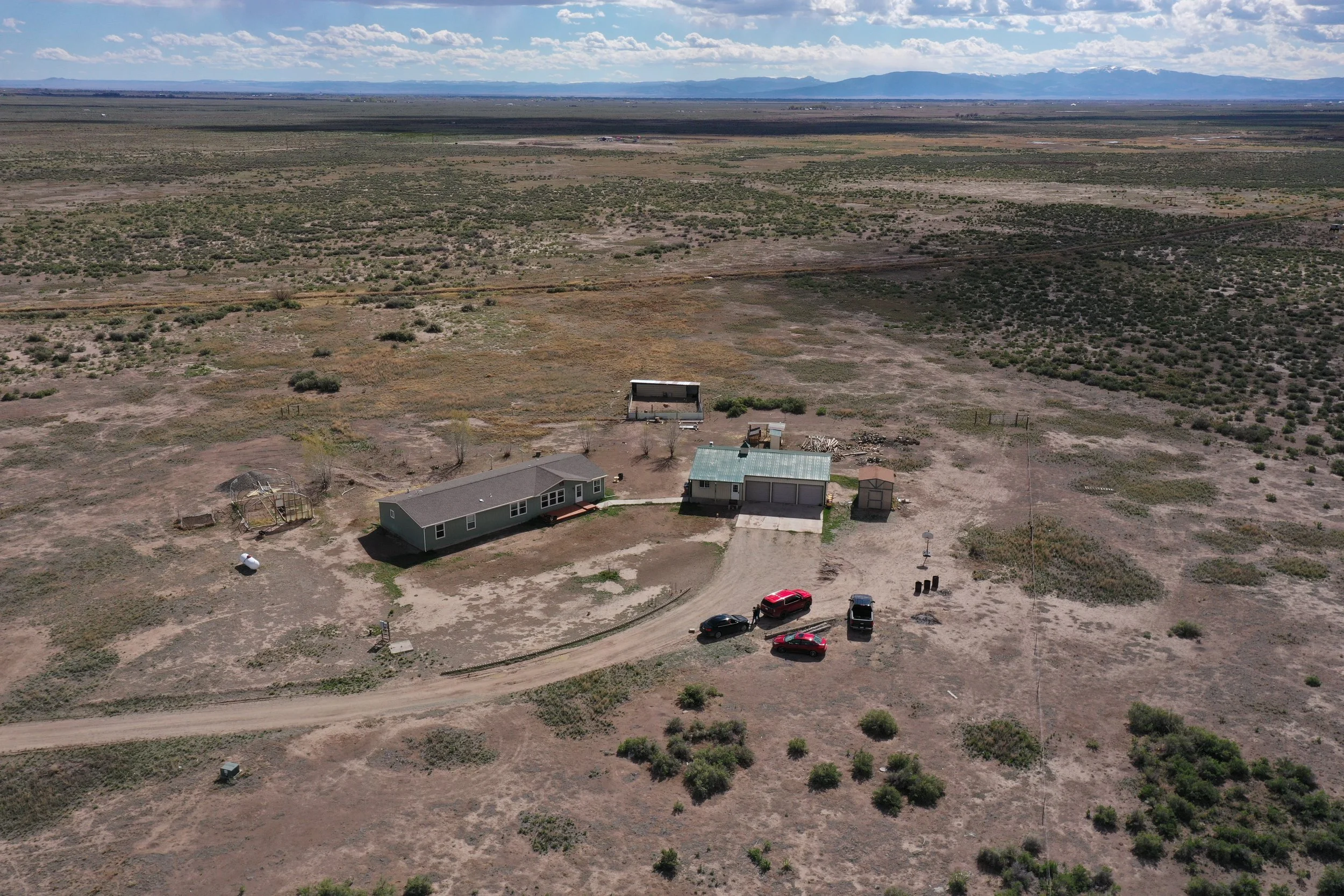 Aerial view of a rural area with three houses, cars, and open land, with mountains in the distance.