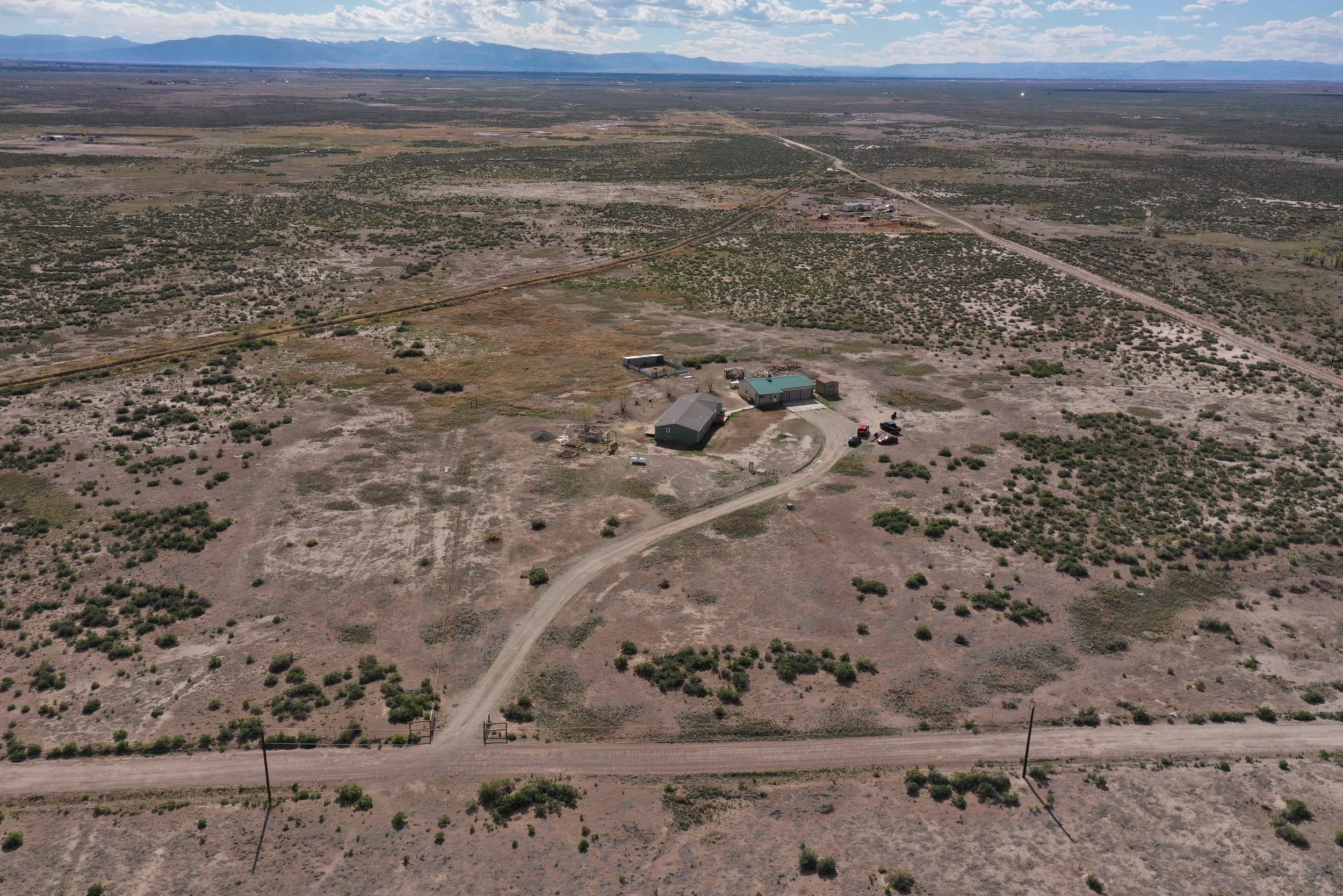Aerial view of a rural desert landscape with sparse vegetation, a few houses, dirt roads, and distant mountains under a partly cloudy sky.