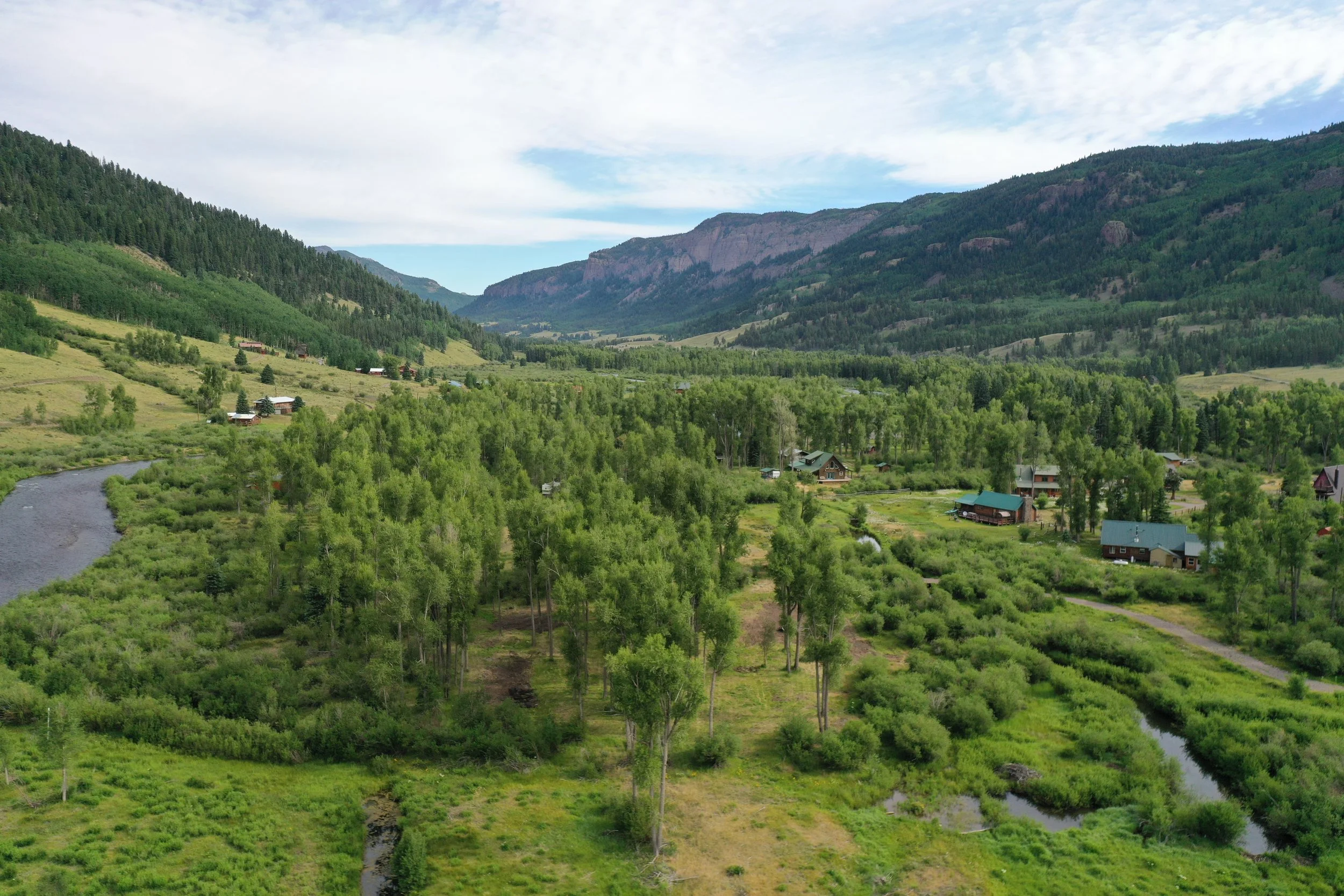 A green landscape with rolling hills, a river, scattered houses, trees, and mountains in the background under a partly cloudy sky.