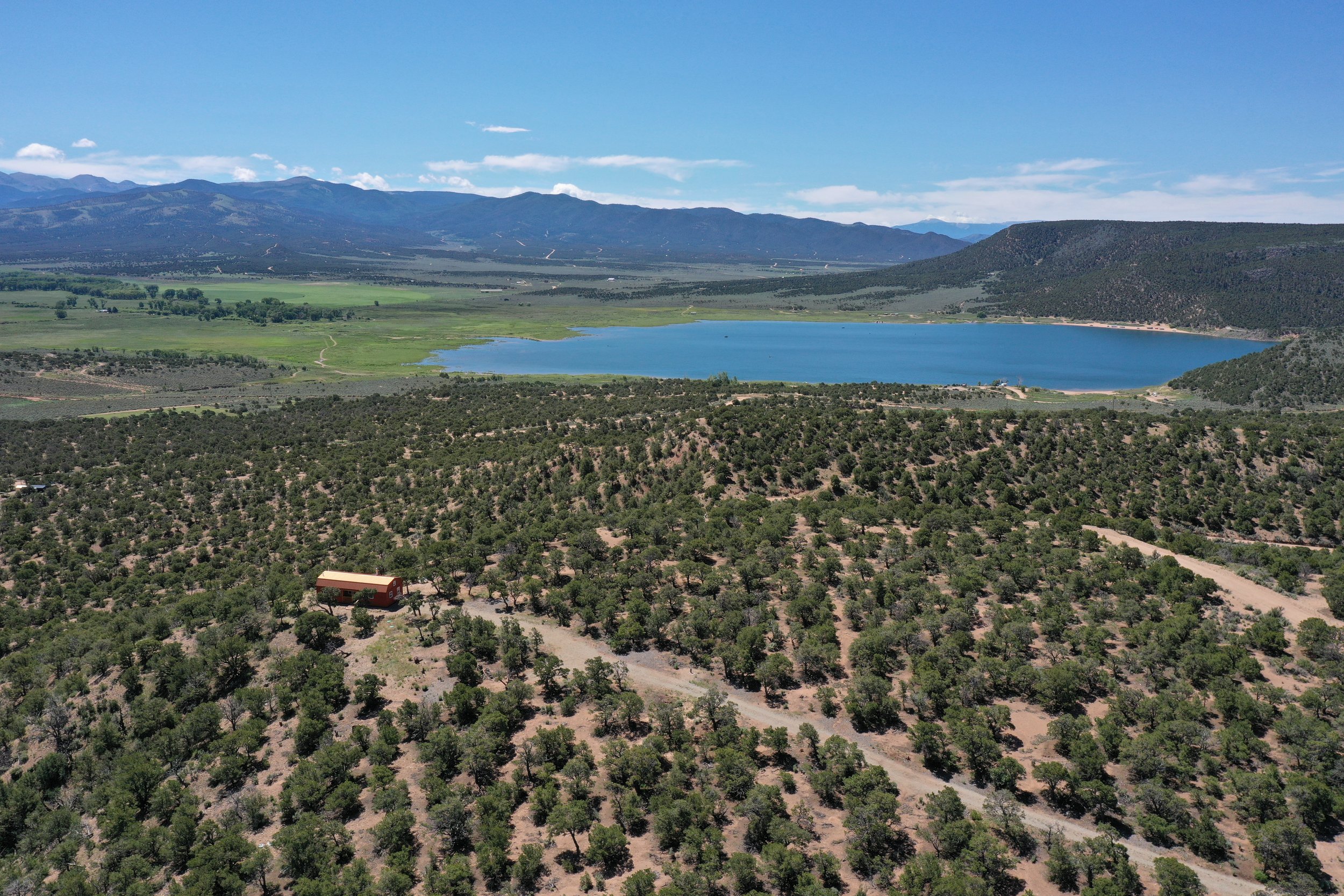 A scenic landscape of a large lake surrounded by green fields, hills, and mountains under a bright blue sky with scattered clouds.