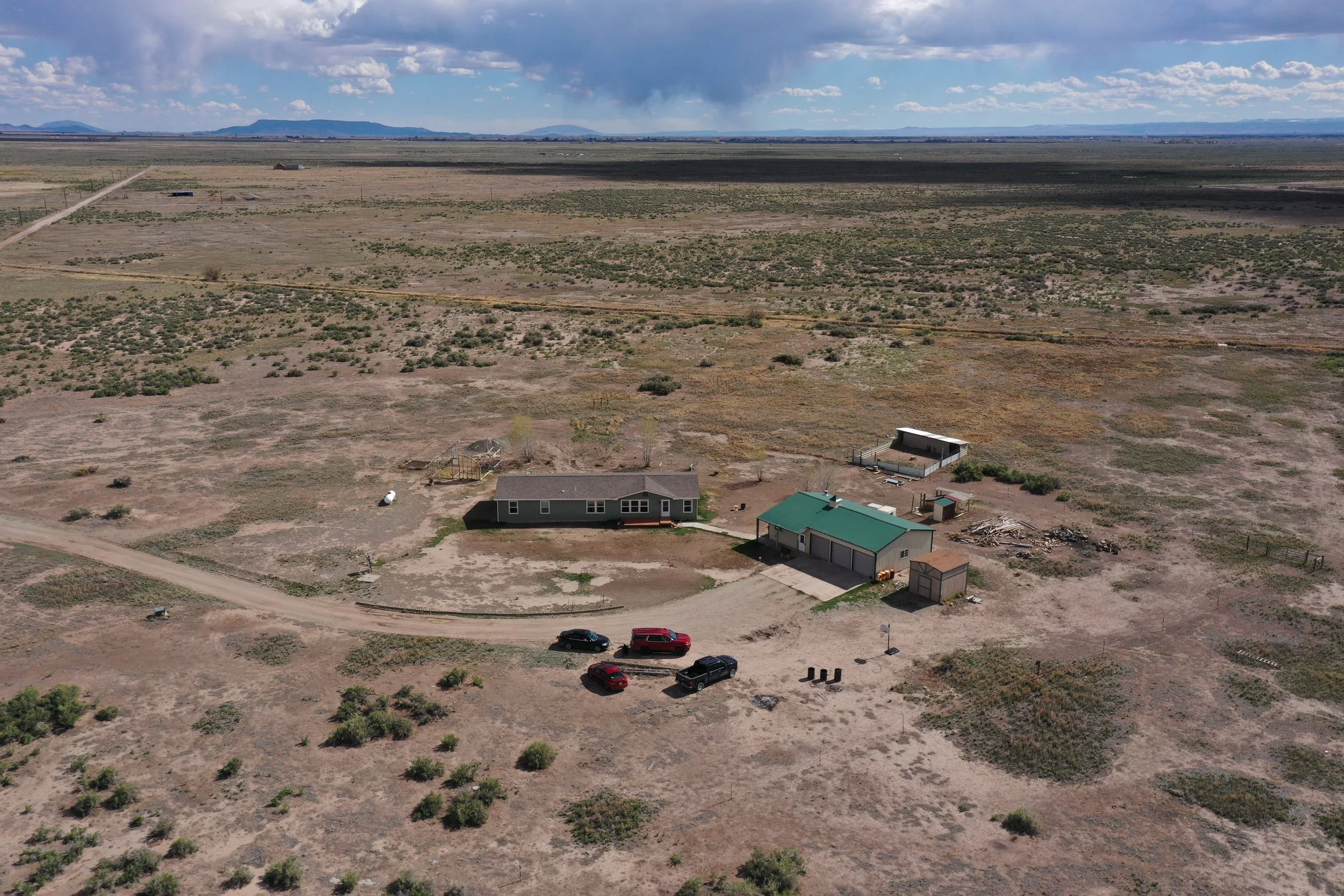 An aerial view of a rural desert landscape with a few houses and cars, sparse vegetation, and distant mountains under a partly cloudy sky.