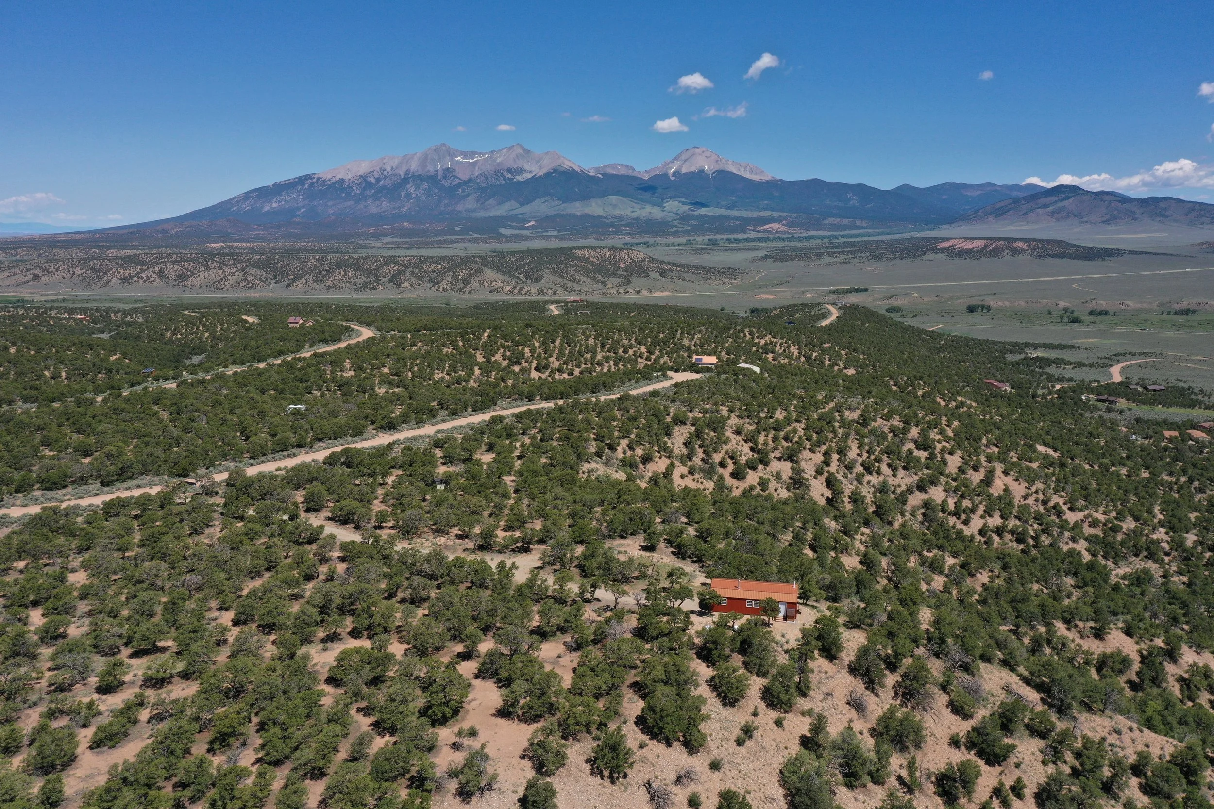 Aerial view of a mountainous landscape with a green forest, dirt roads, and houses in the foreground, and a large mountain range under a blue sky with some clouds.