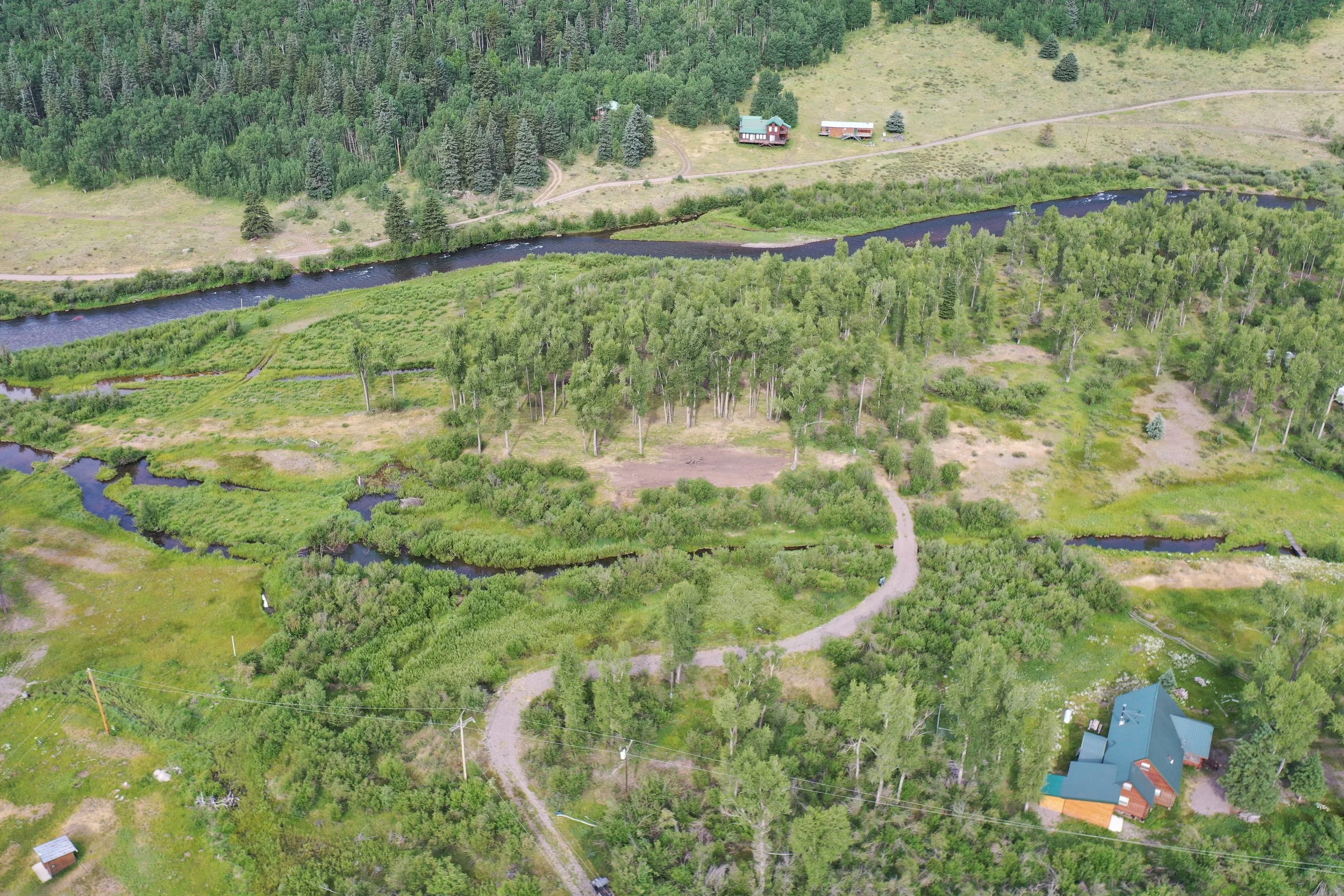 An aerial view of a rural landscape with a winding river, green trees, a dirt road, and several buildings, including houses and small structures.