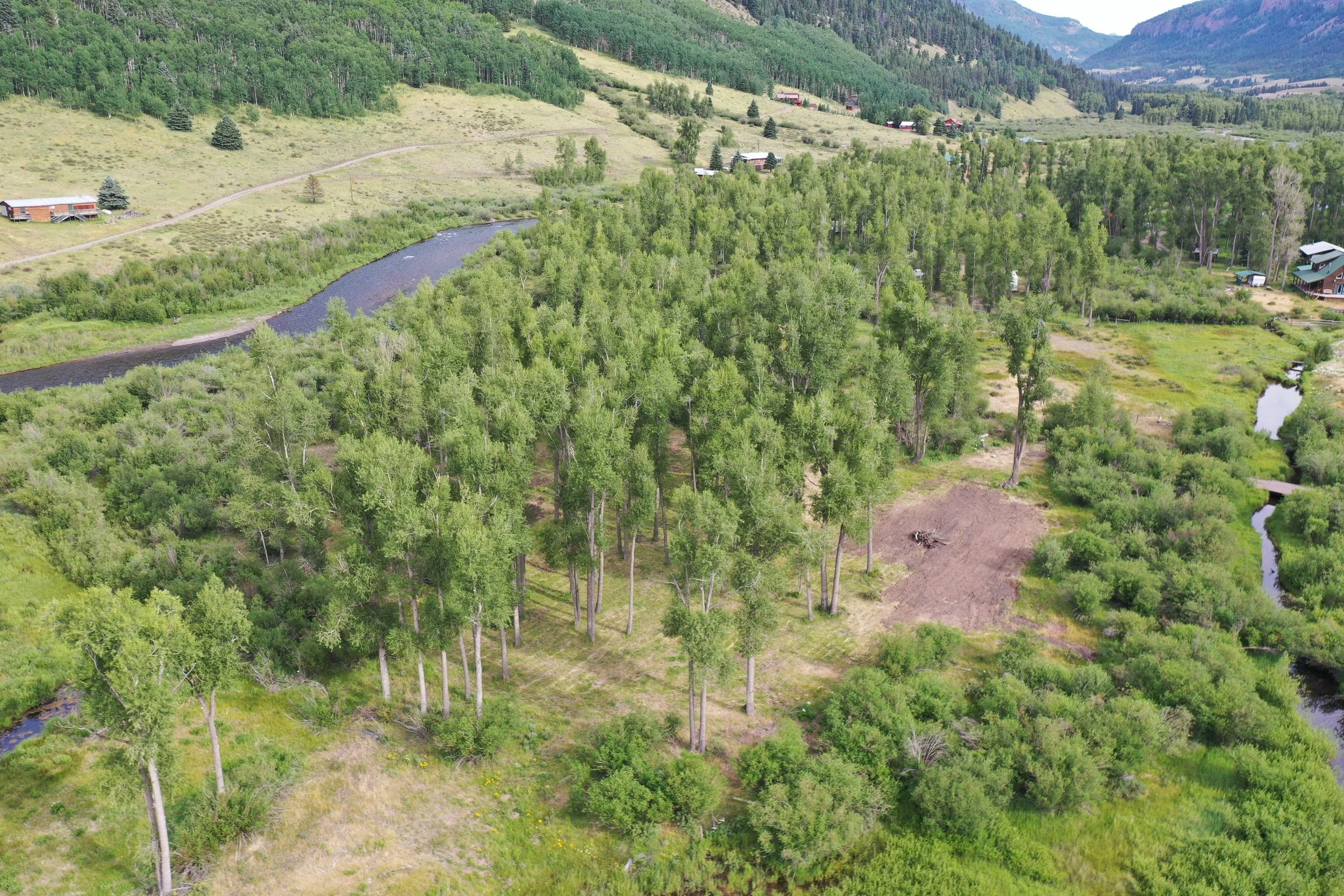 A scenic aerial view of a lush green rural landscape with a river meandering through the forest, scattered houses, and rolling hills in the background.