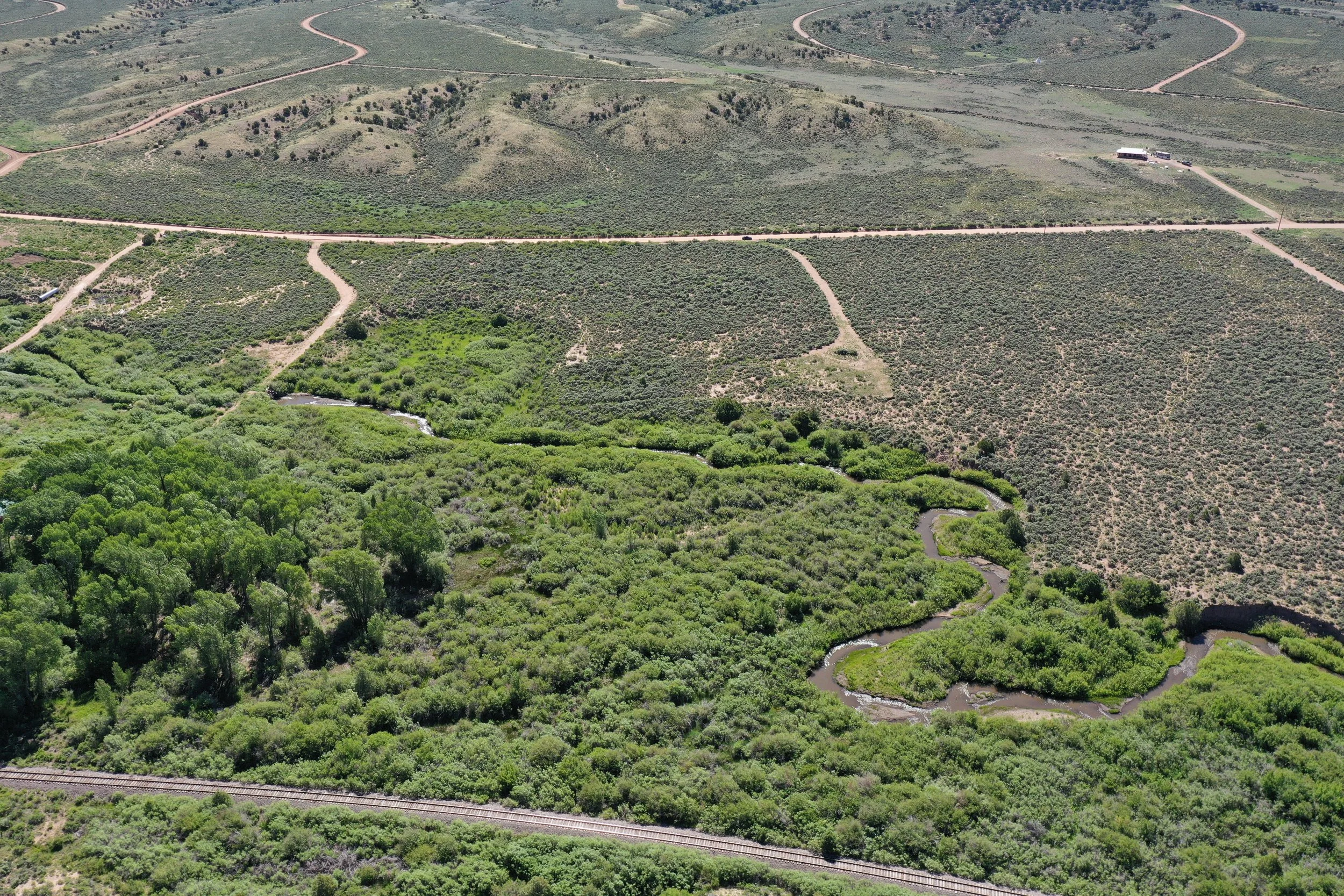 Aerial view of a vast landscape featuring a winding river surrounded by dense green trees, dirt roads crisscrossing through a hilly area with some sparse vegetation, under clear skies.