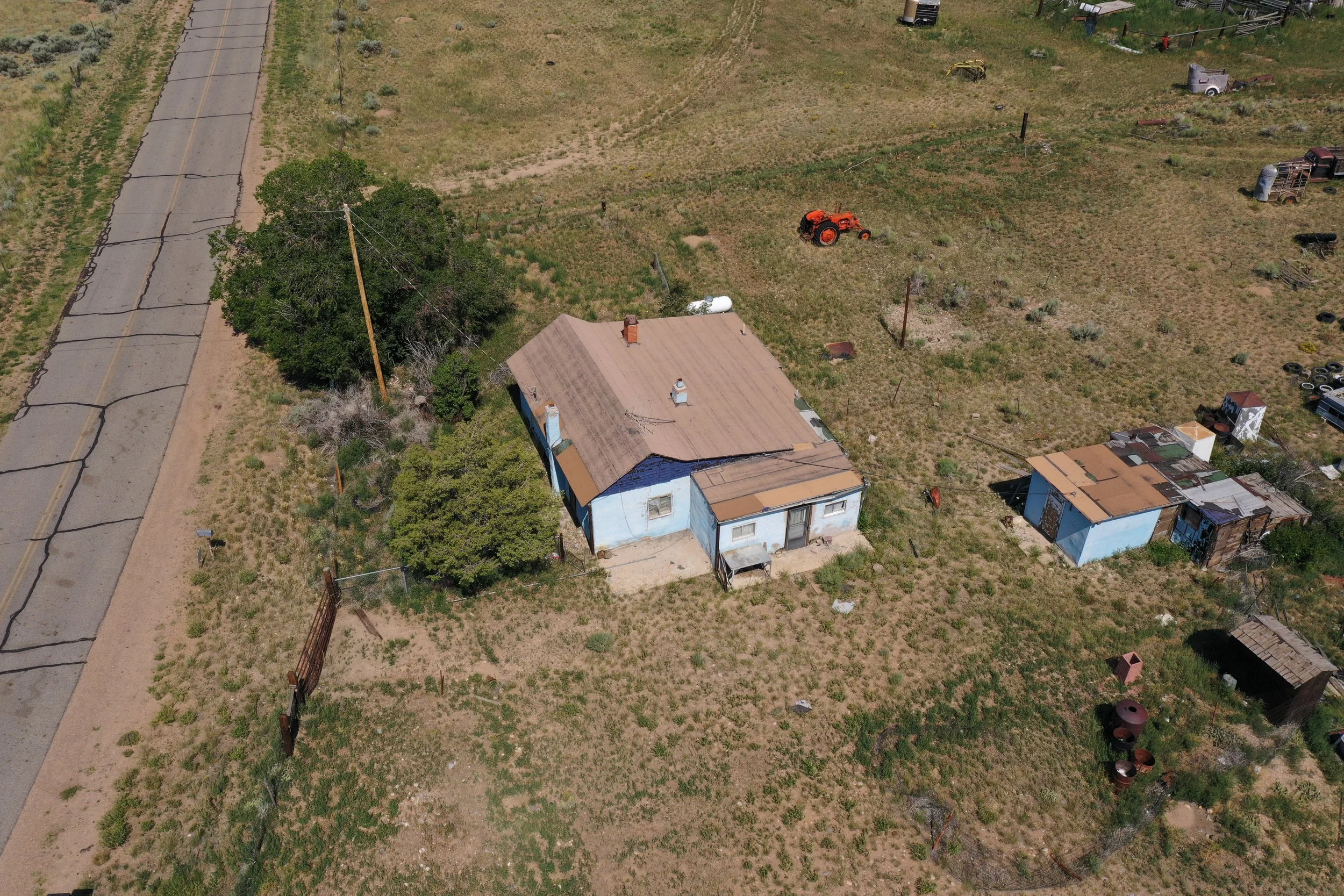 Aerial view of a rural area with a house, a tractor, a tree, and various farm equipment and debris, along a cracked asphalt road.