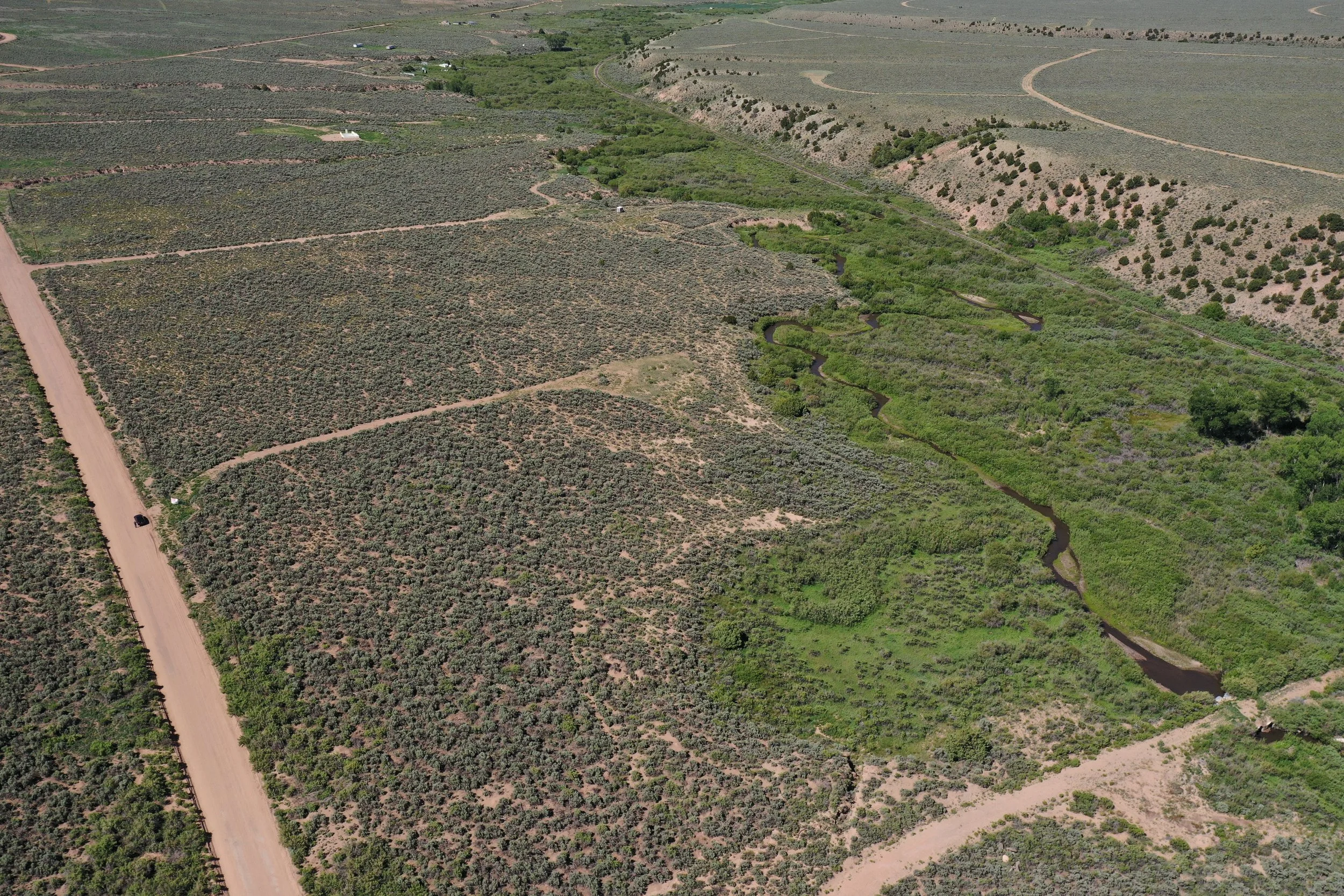 Aerial view of a vast rural landscape with patches of green bushes and trees, winding roads, and a small creek running through the land. There are some scattered structures and a dirt road on the bottom left corner.