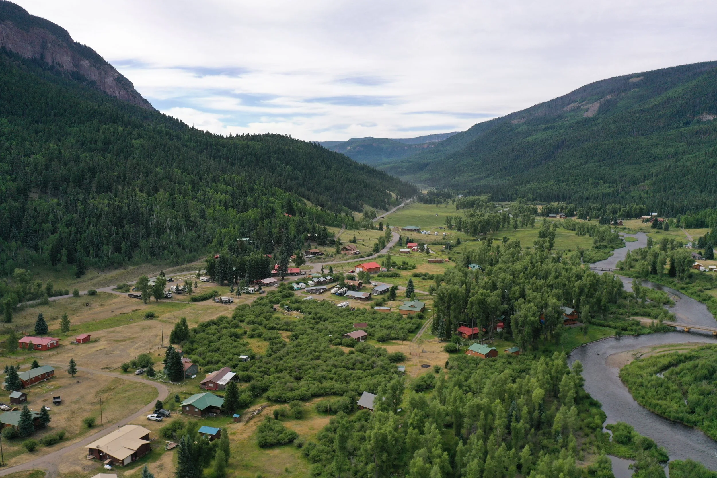 Aerial view of a lush green valley with scattered houses, a winding river, and surrounded by dense forested mountains under a cloudy sky.