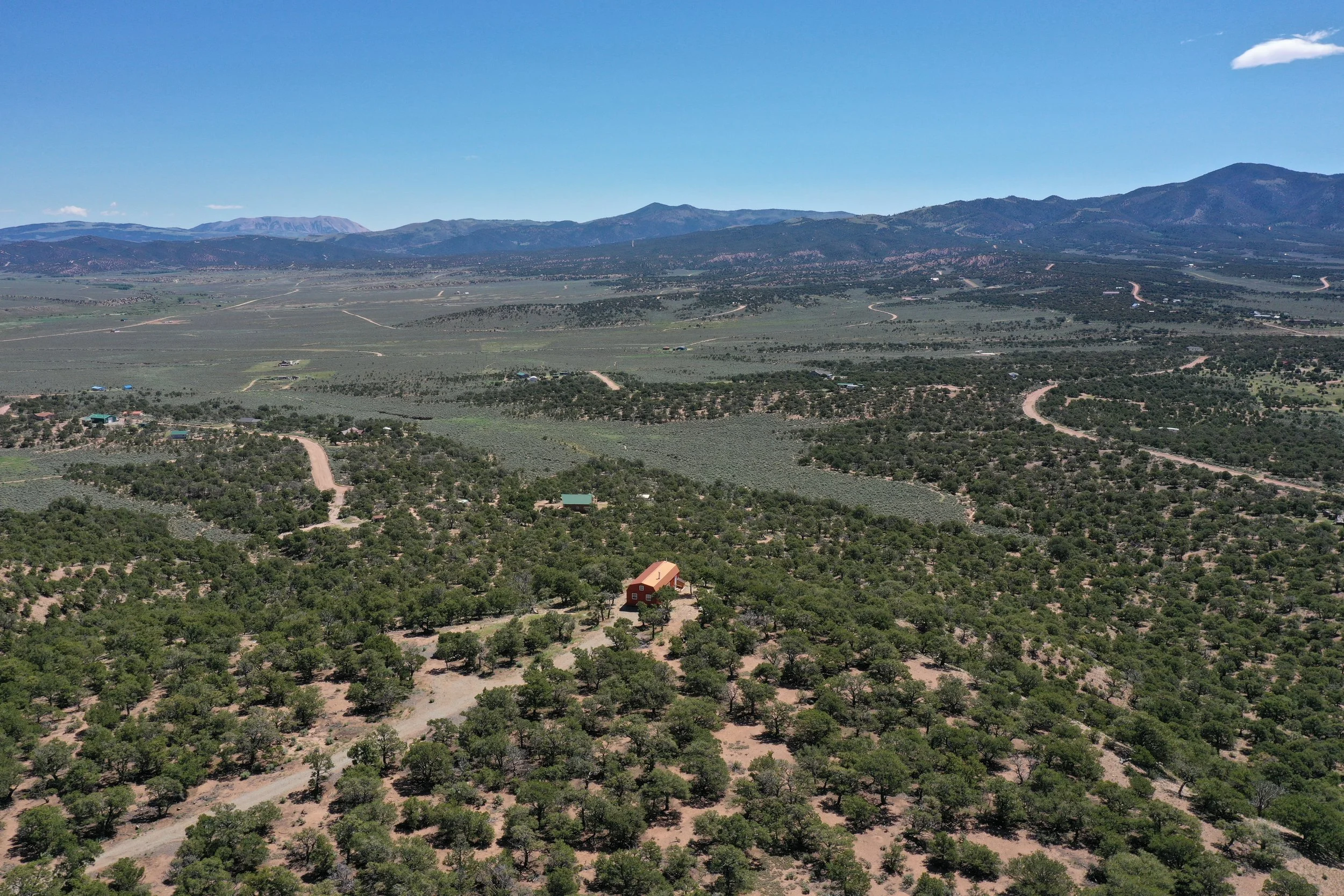 A wide view of a rural landscape with scattered trees, a few houses, and winding dirt roads set against a backdrop of mountains under a clear blue sky.