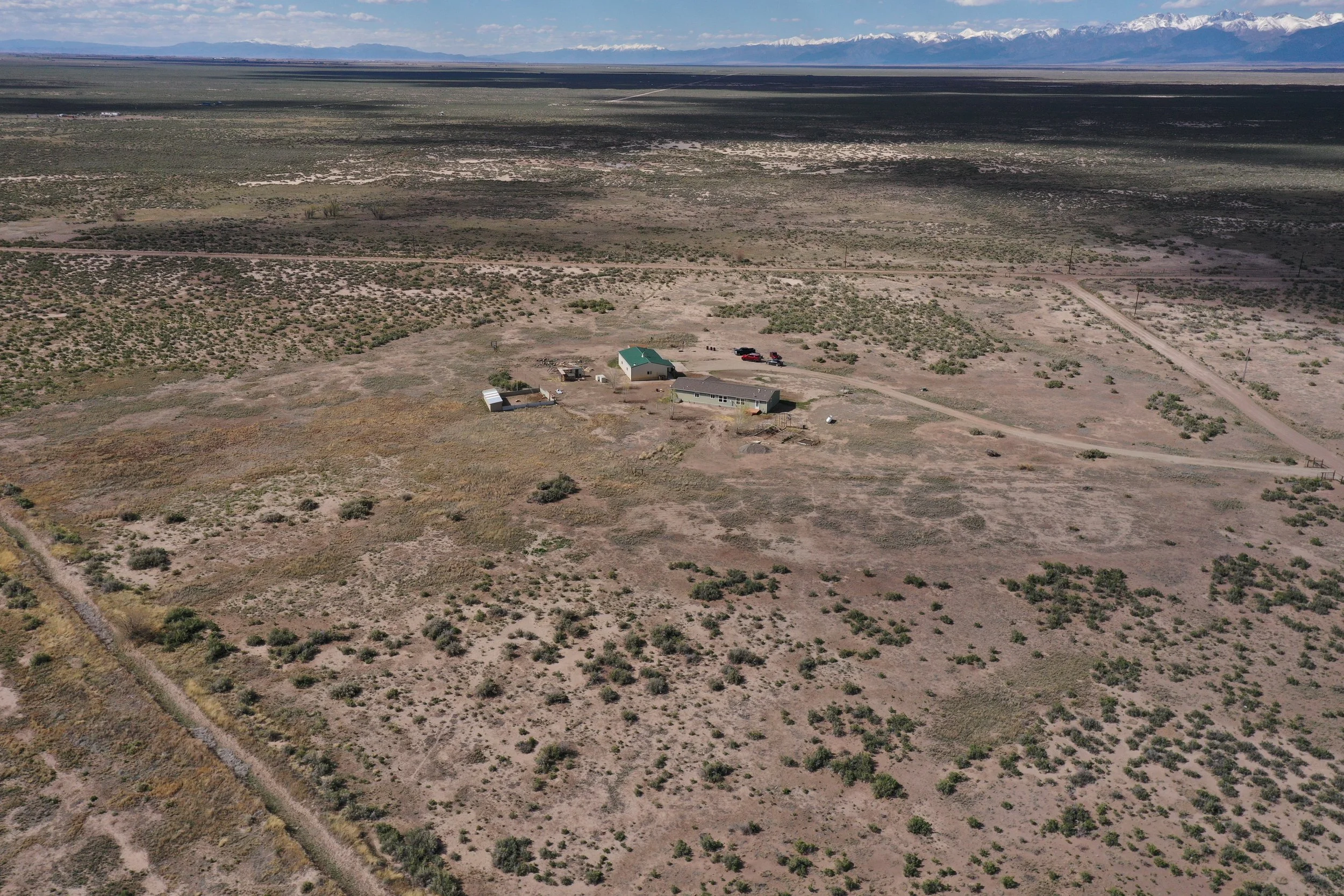 Aerial view of a rural desert landscape with scattered bushes, two small houses, and a few vehicles, with distant snow-capped mountains under a partly cloudy sky.
