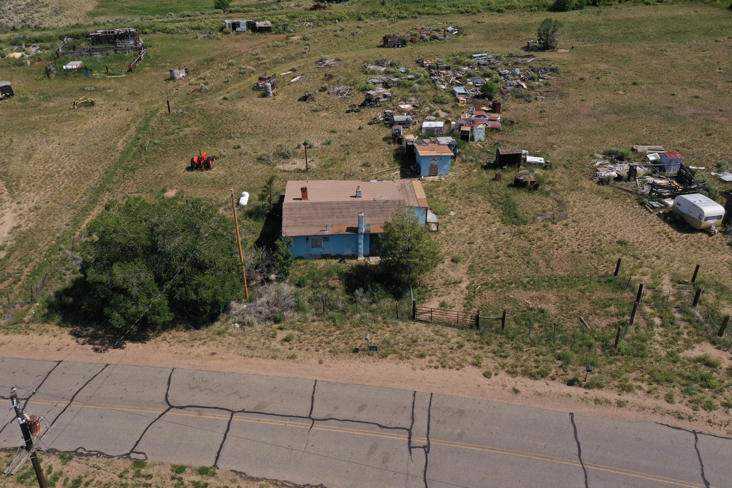 Aerial view of a rural area with a small house surrounded by an open field scattered with abandoned vehicles, debris, and old structures. A paved road runs along the bottom of the image with power lines overhead.