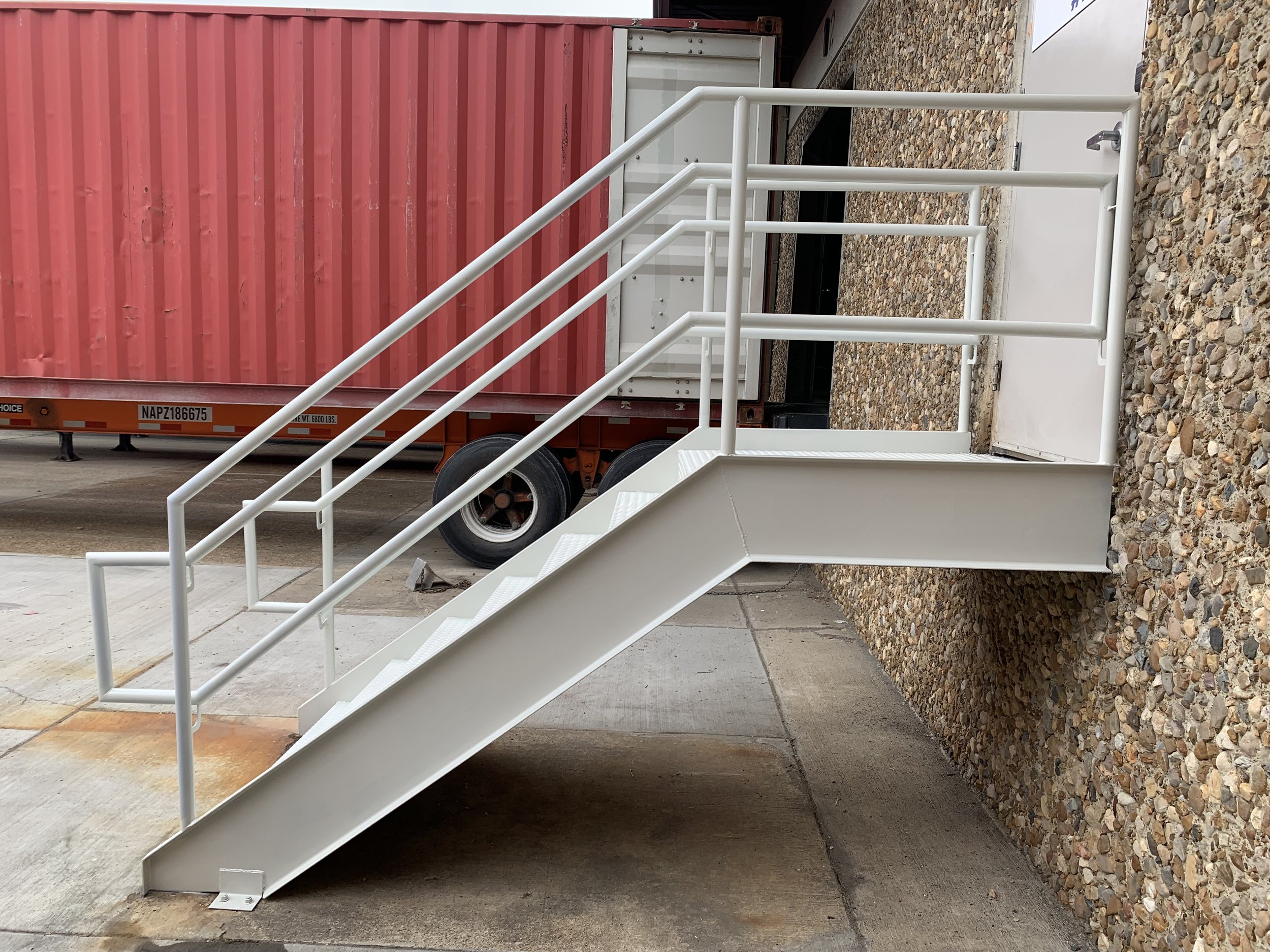 White metal exterior staircase with handrails leading to a door at the top, placed on a concrete and gravel surface, with a red shipping container and truck in the background.