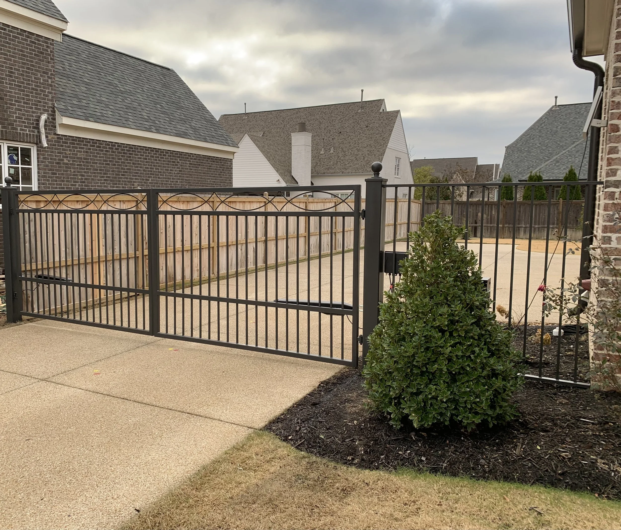 Residential backyard with a black metal fence and gate, a small shrub, a concrete patio, and neighboring houses with landscaped yards under an overcast sky.