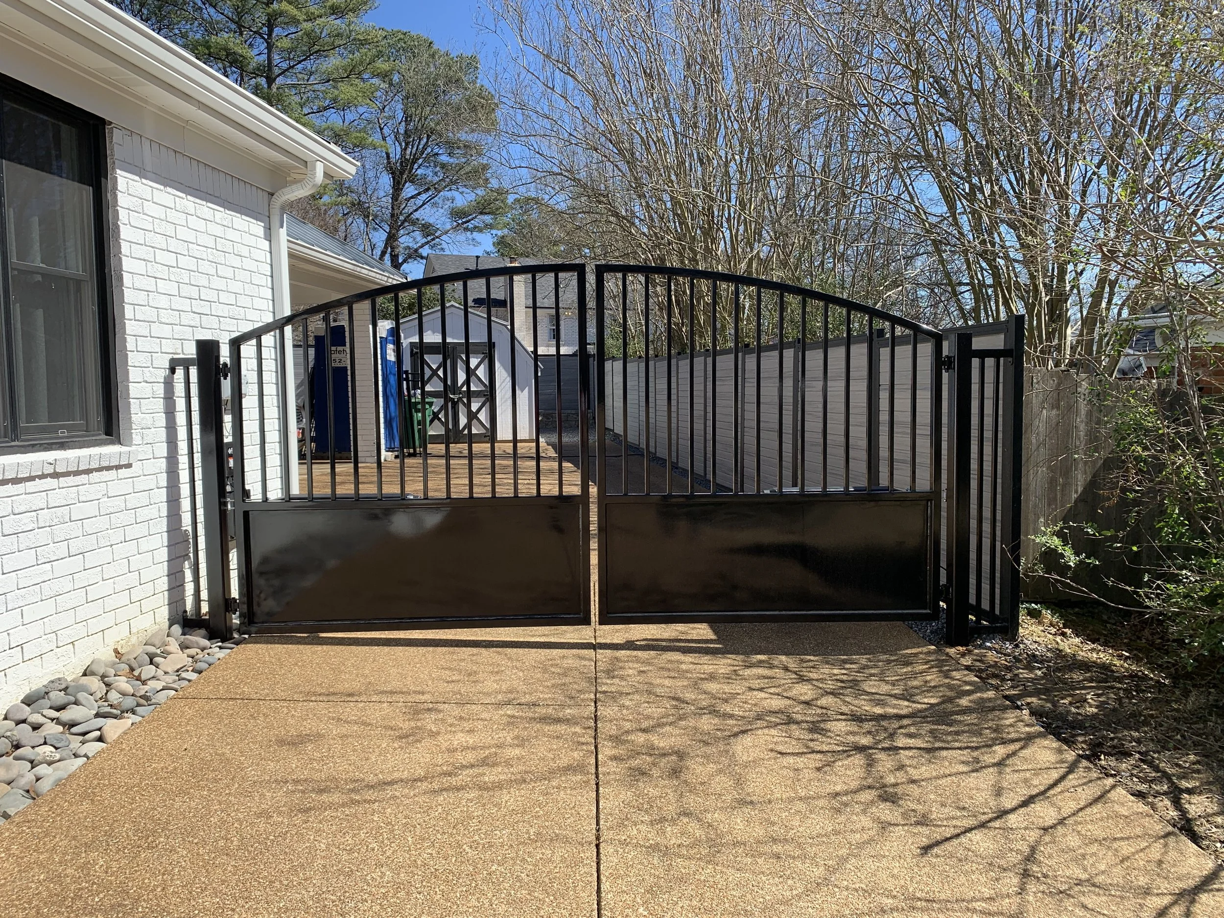 Black metal gate on a concrete driveway leading to a backyard with wooden deck, shed, and trees.