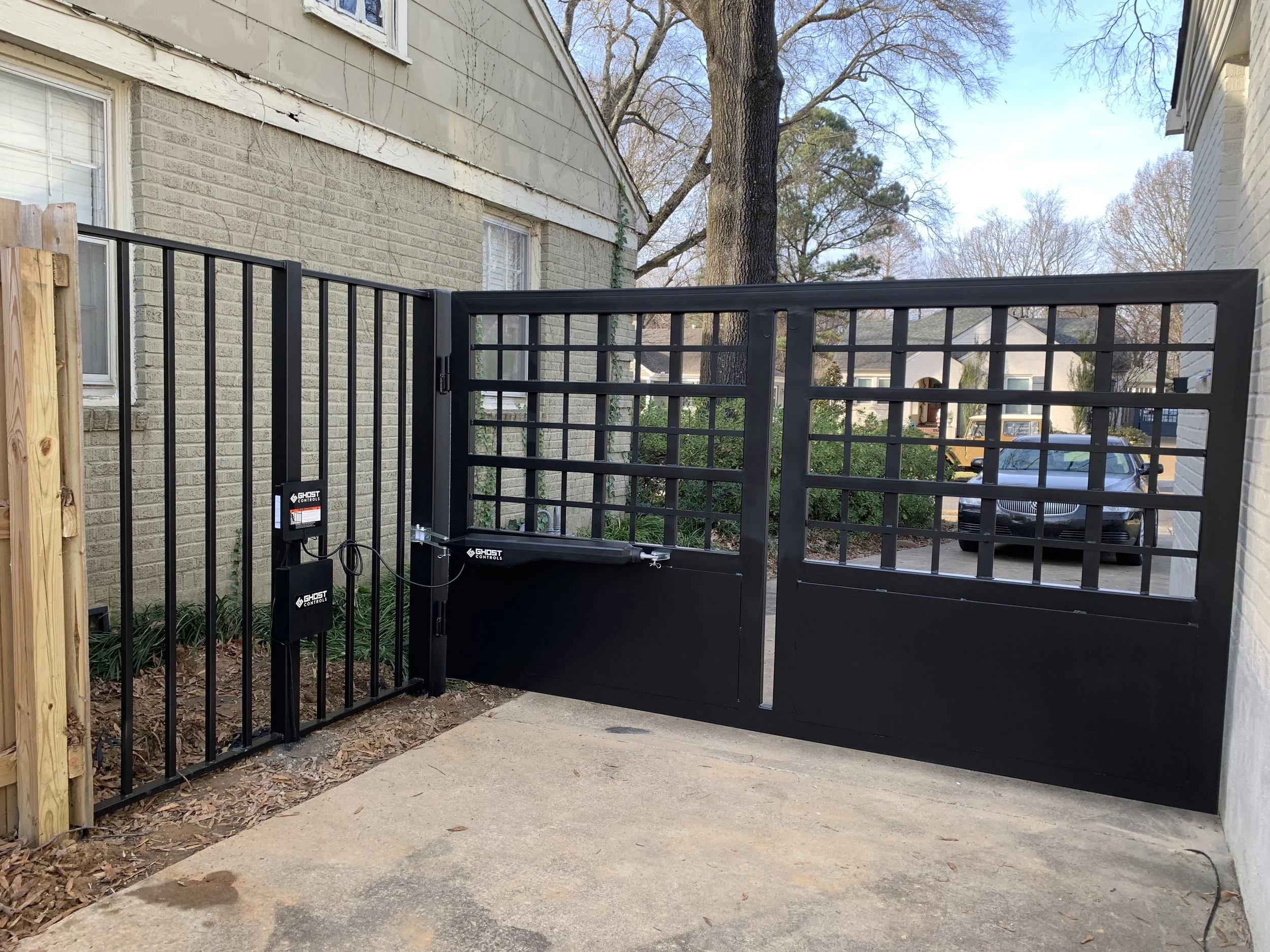 Black automated gate with a grid pattern, installed between two residential buildings, with an electronic control box mounted on a fence post.