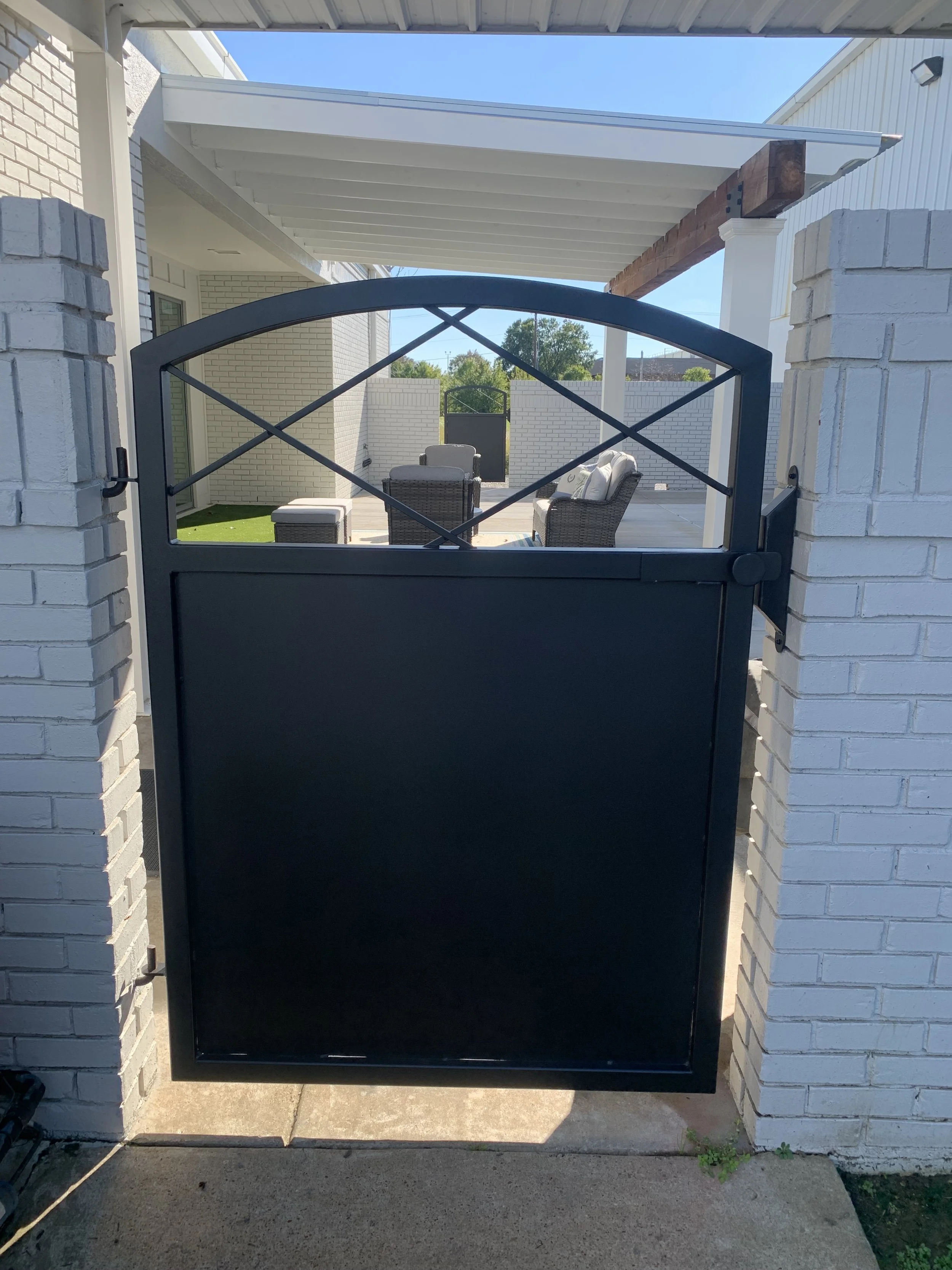 Black metal gate open to a backyard patio with outdoor furniture under a white pergola, surrounded by white brick walls.