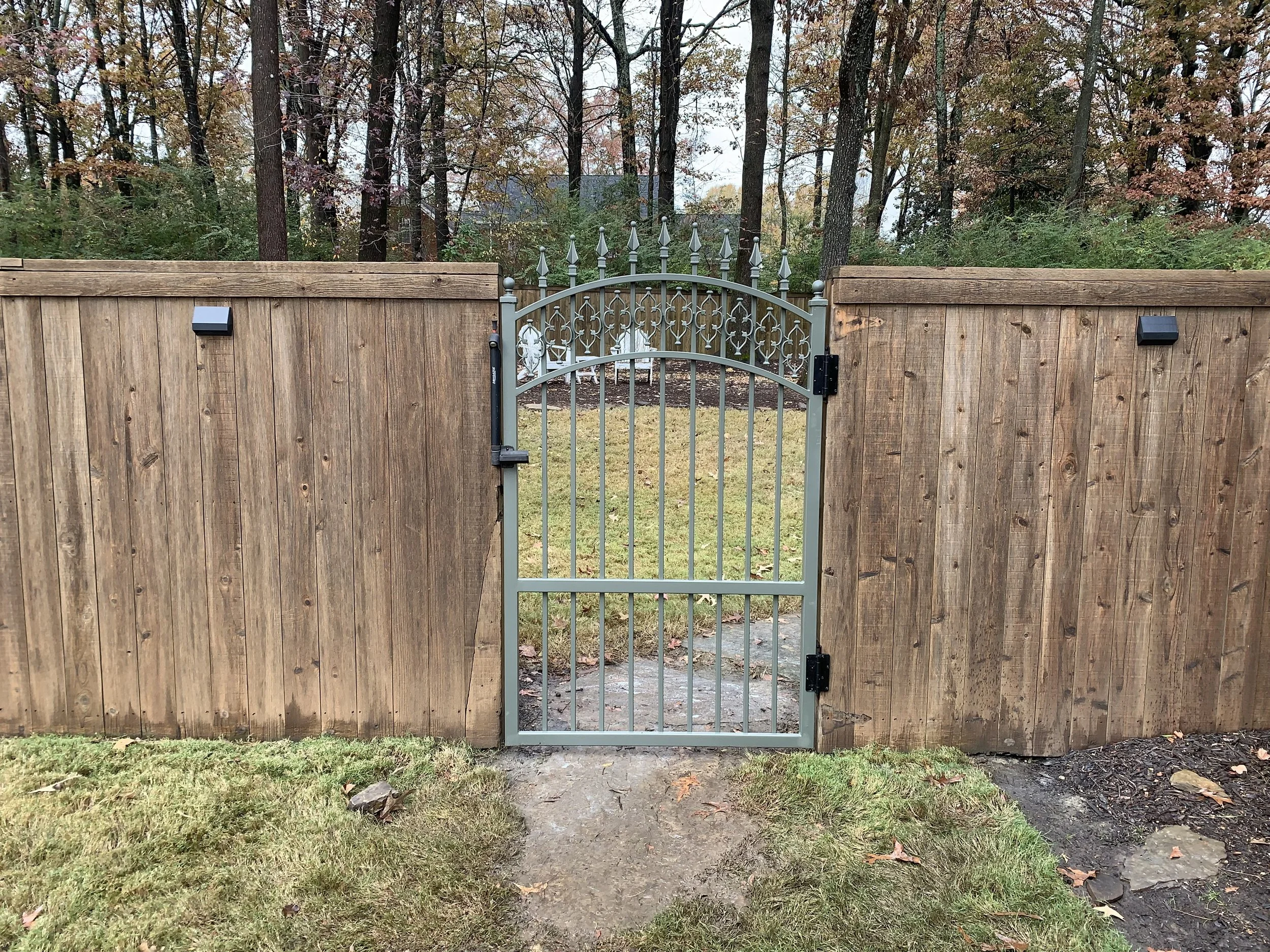 A metal gate with decorative spikes, set between two wooden fences, opens onto a grassy yard with trees in the background.