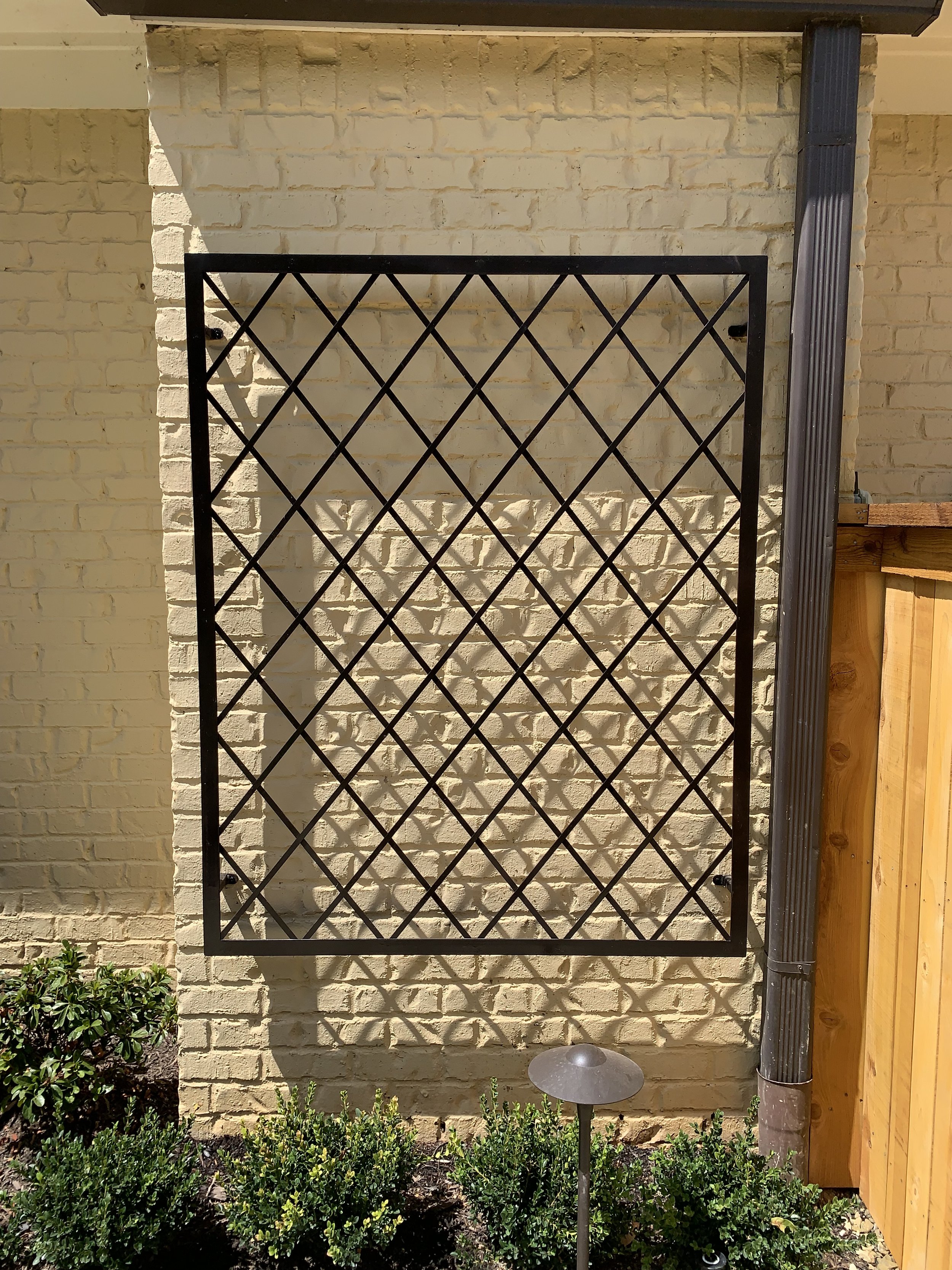 Black metal trellis mounted on a light yellow brick wall, with a garden bed of green bushes and a small outdoor light in front.