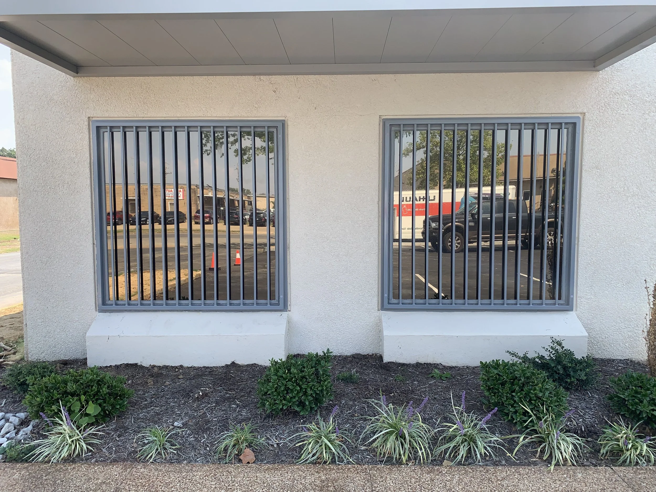 Exterior of a building with two large windows covered by metal bars, and a small landscaped area with bushes and plants in front.