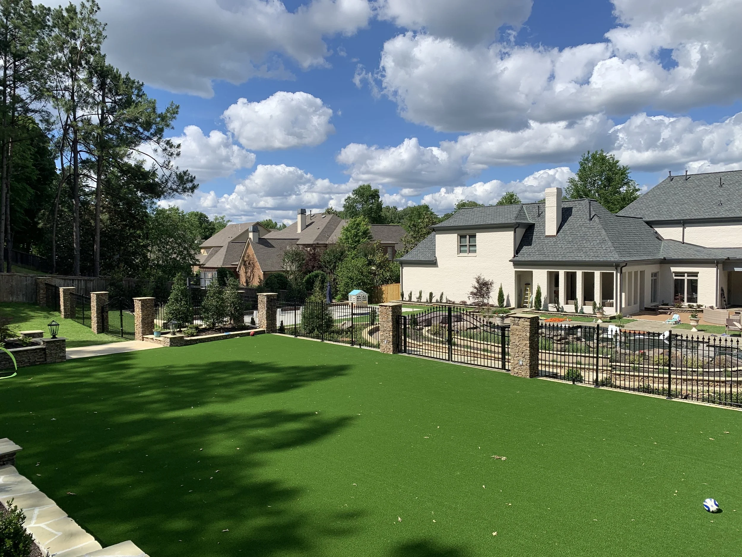 A backyard with artificial grass, a stone-paved patio, and a black metal fence, with a modern white house and neighboring houses in the background under a partly cloudy sky.