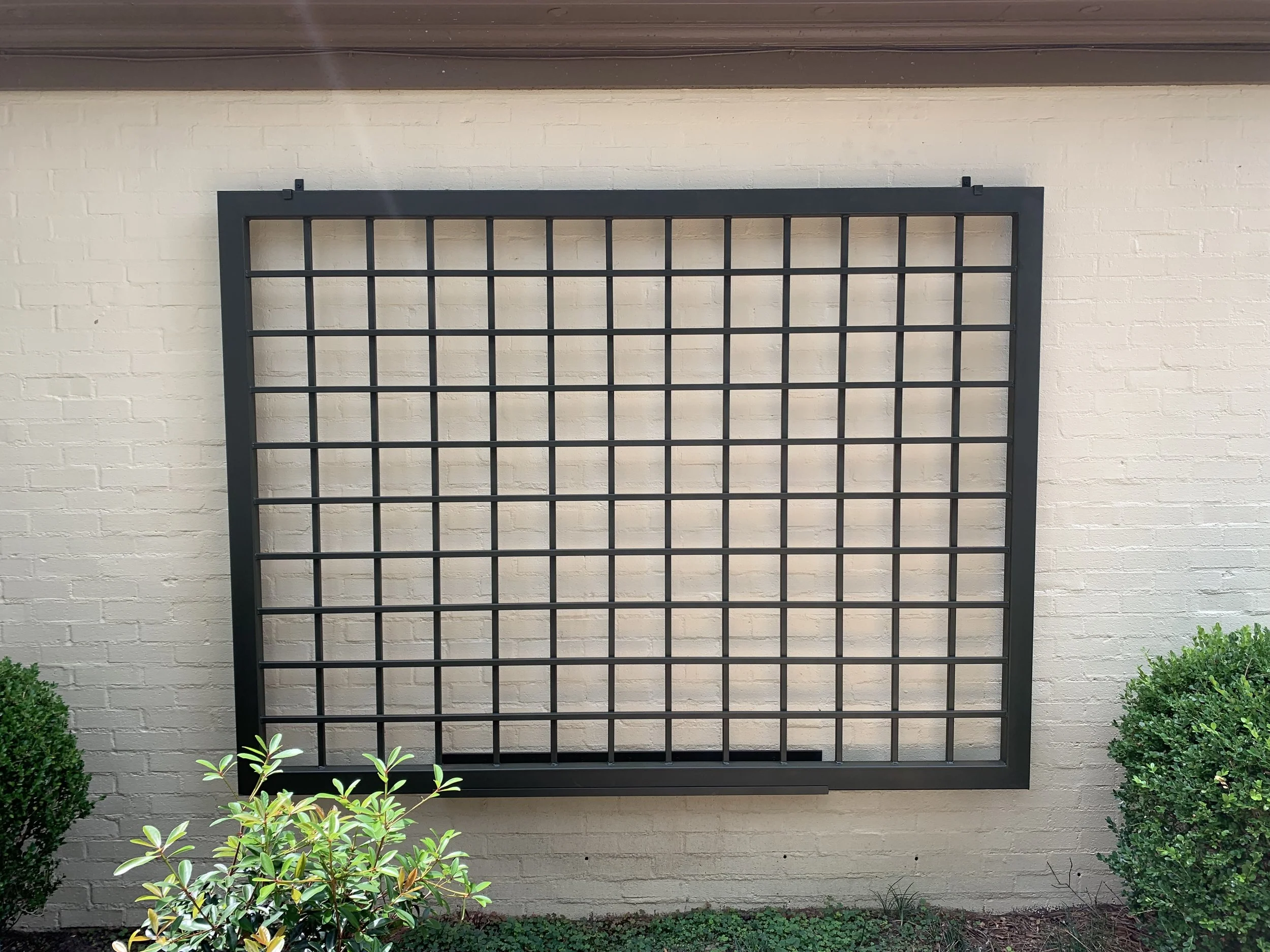 Empty black grid frame mounted on a white brick wall, with bushes at the bottom. Trellis