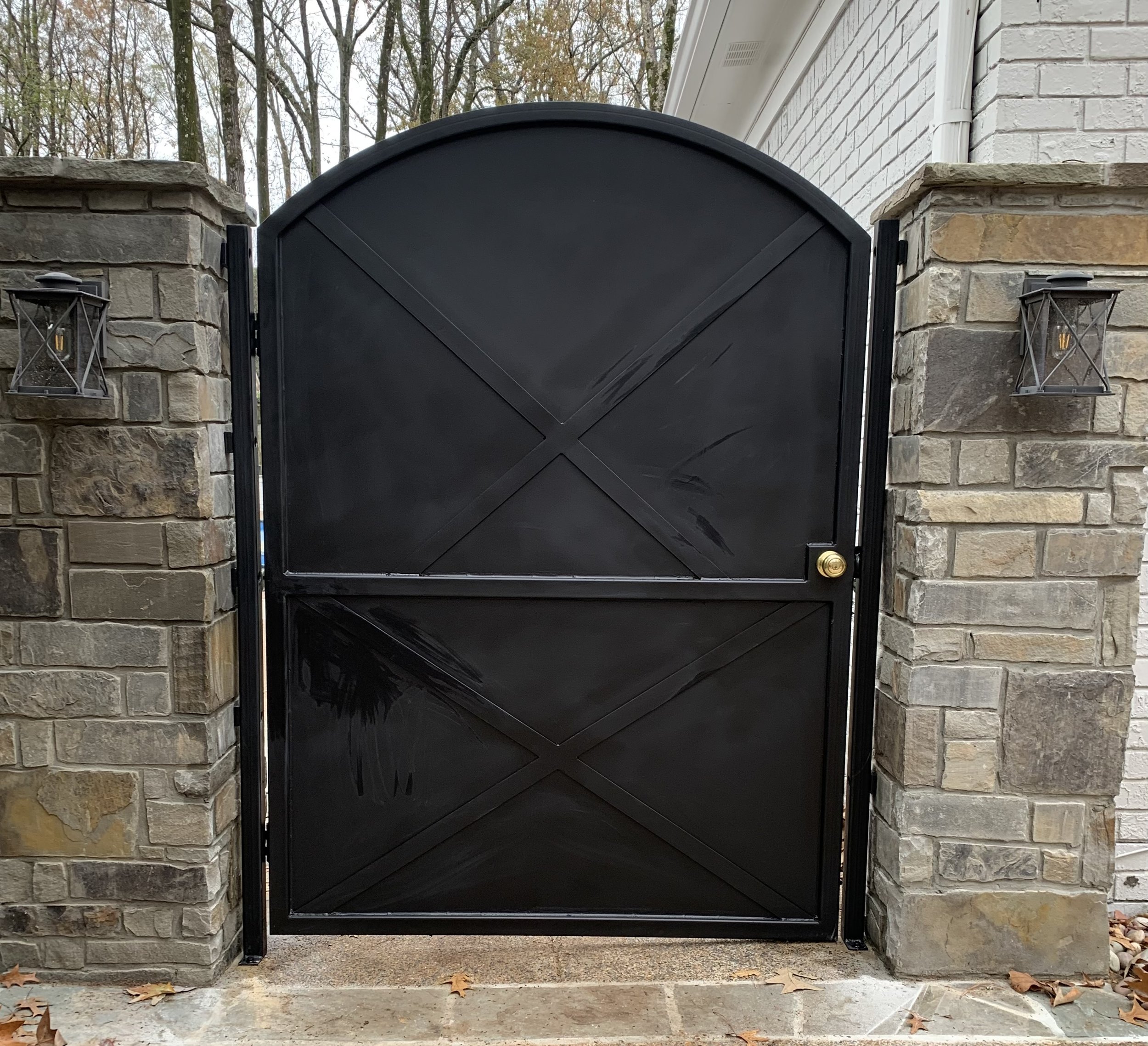 Black metal gate with a rounded top, mounted between two stone pillars, with wall-mounted lanterns on each side